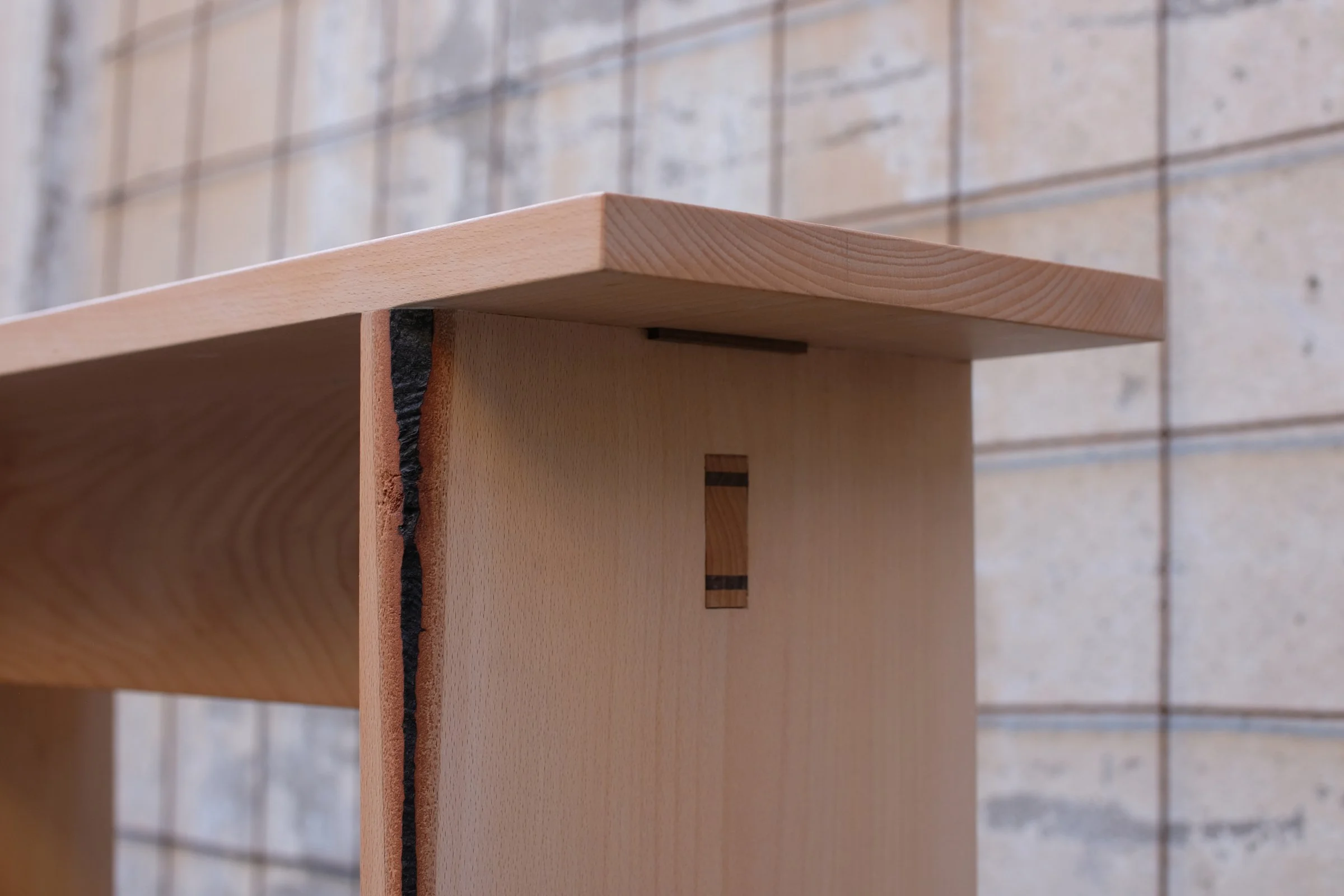 Close-up of a wooden furniture piece, showing a wedged through-tenon joint with dark colored wedges, against a textured stone background.