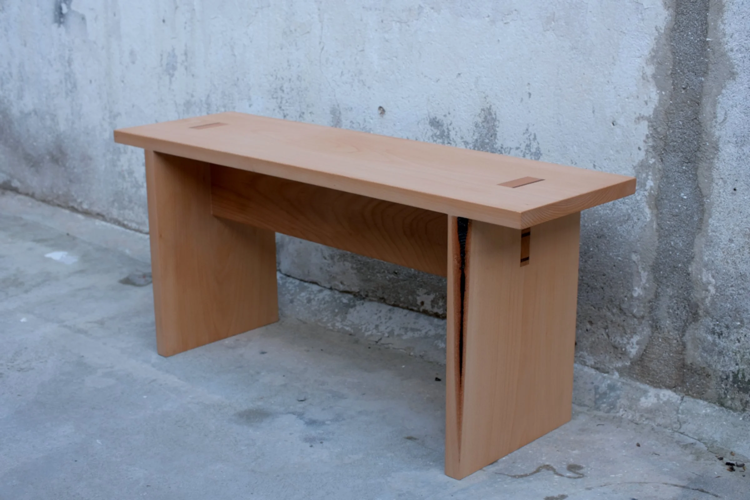 A minimalist wooden bench with wedged through-tenons and horizontal support, positioned against a textured concrete wall.