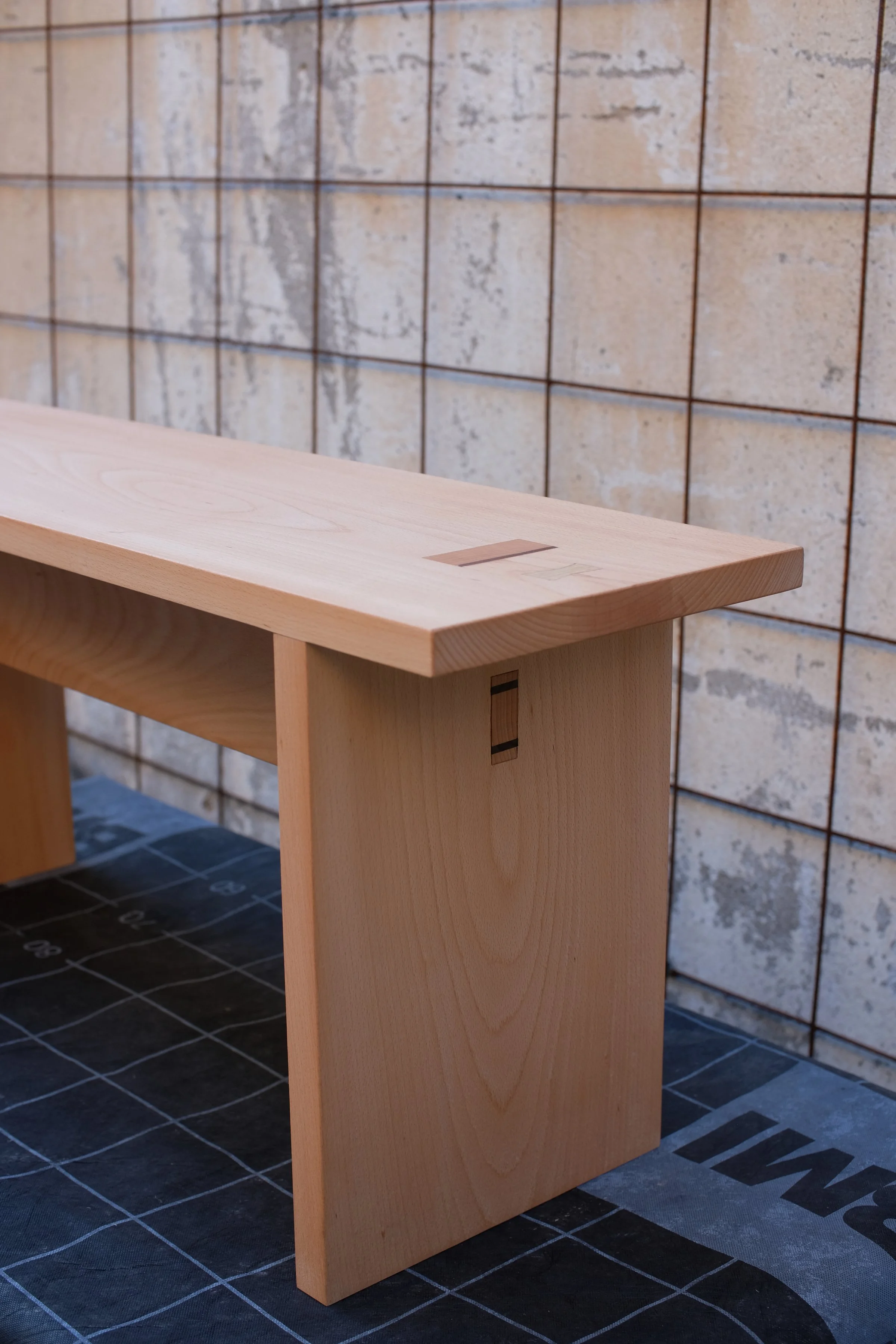 A close-up of a wooden bench with a natural finish against a concrete block wall with a metallic grid.