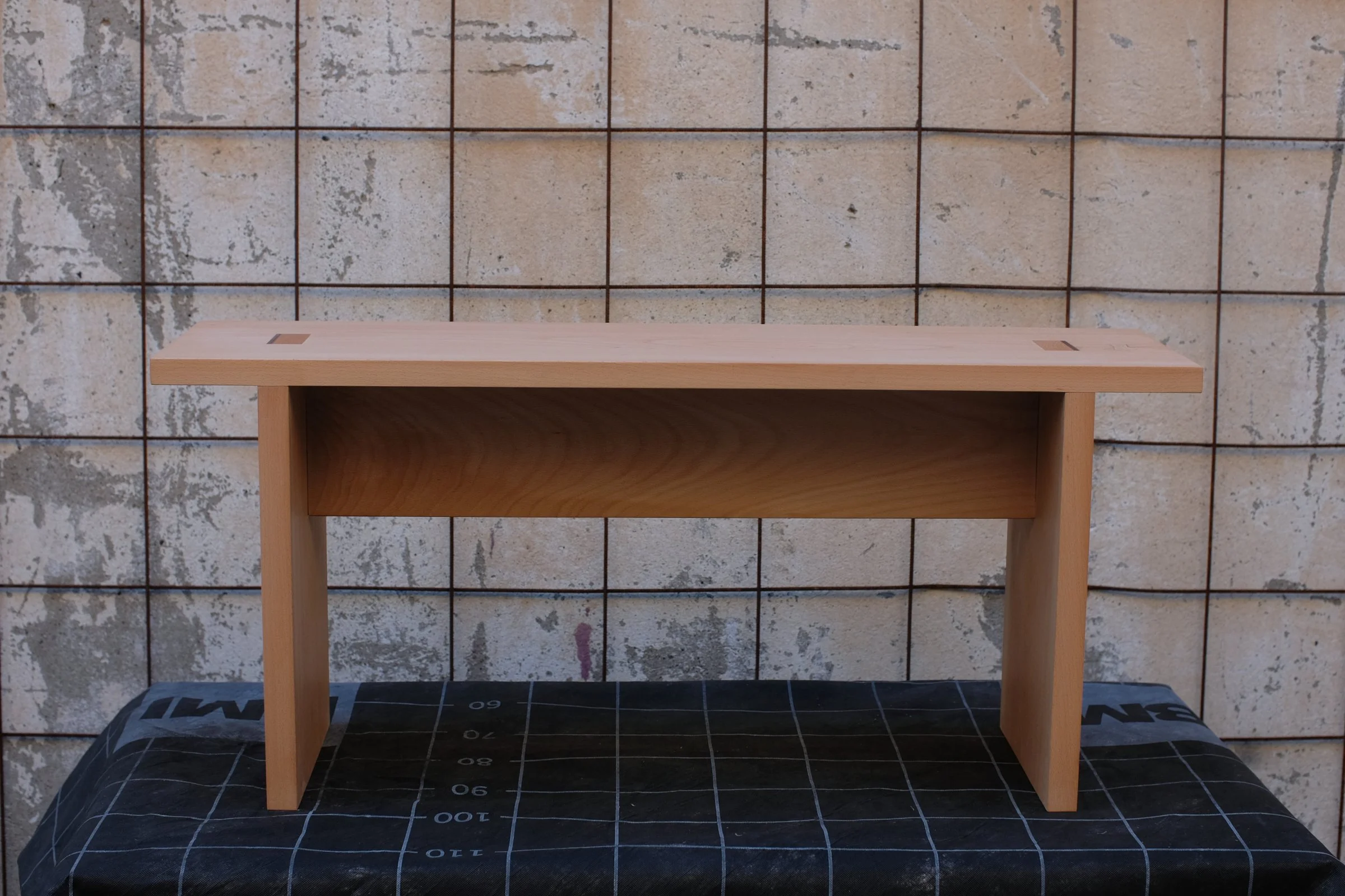 A small, light-colored wooden bench with a simple design, placed on a blac gkrid-patterned surface with measurements, against a textured stone wall background.
