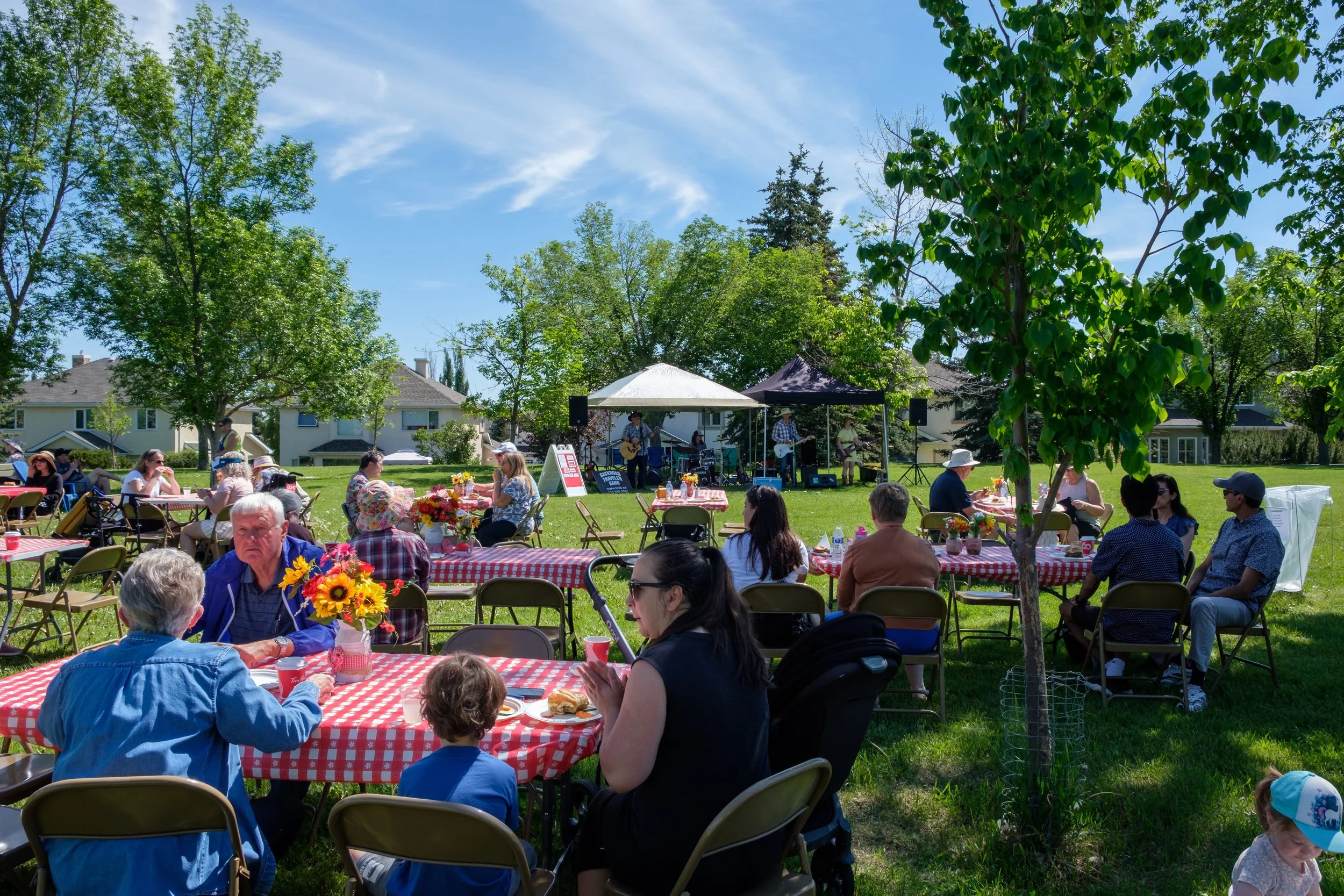 A community outdoor event with people sitting at tables with red and white checkered tablecloths, enjoying food and drinks. In the background, a live band performs under canopies, with trees and houses surrounding the area on a sunny day.