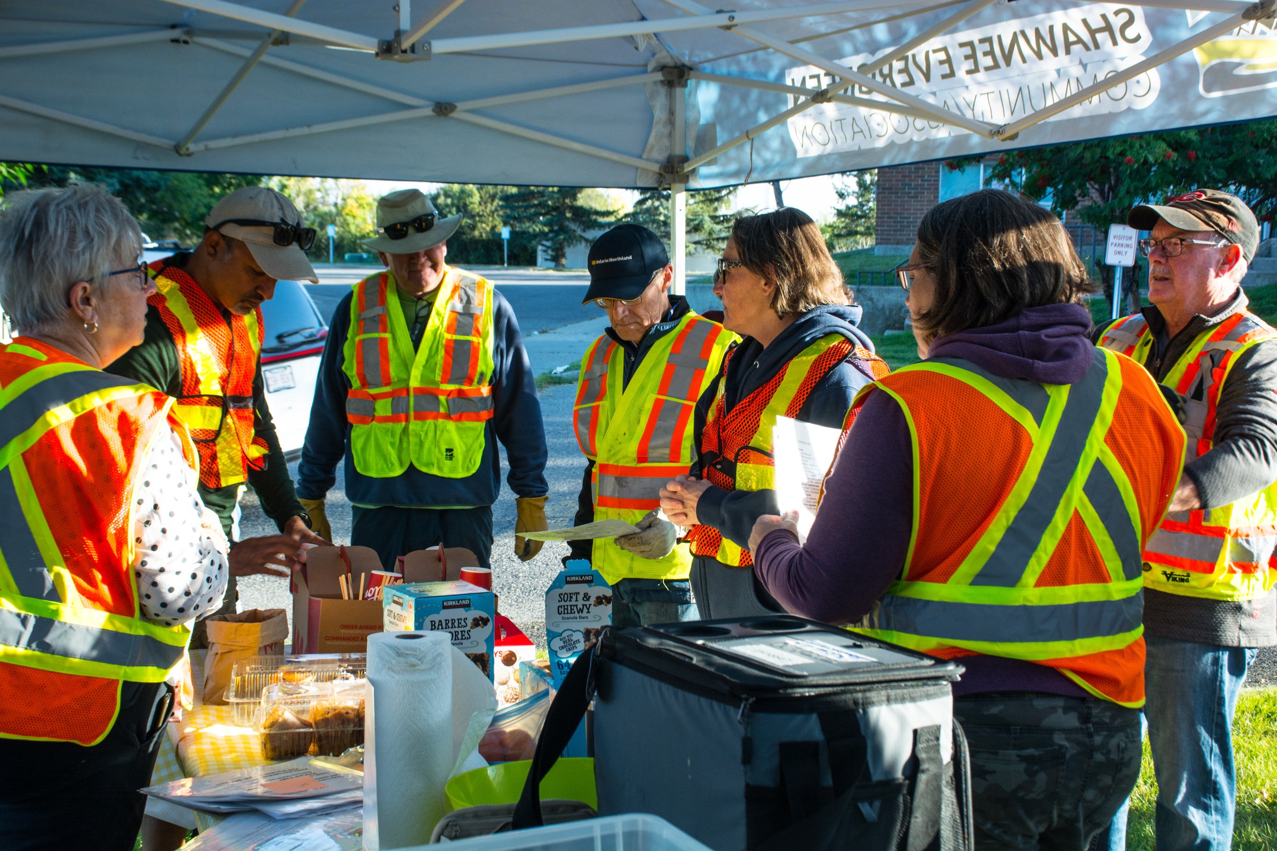 Group of people wearing orange safety vests gathered under a white canopy tent outdoors, with food and drinks on a table.