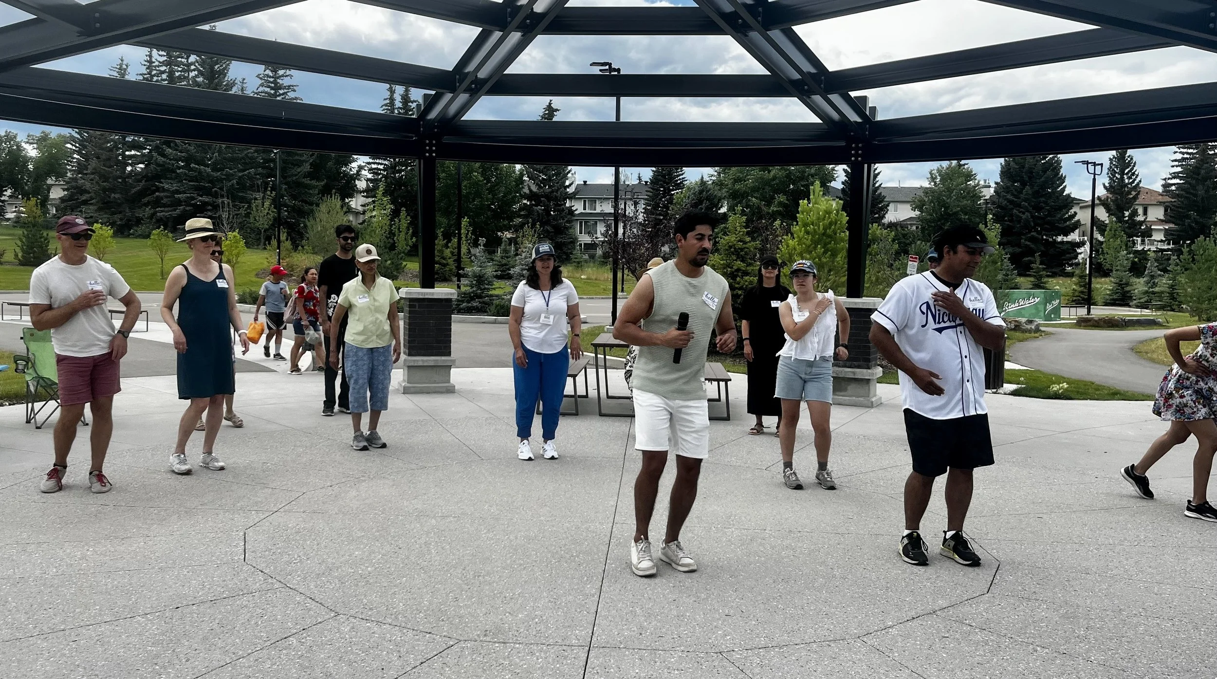 Group of people participating in an outdoor event, some holding microphones and others standing or walking around under a metal gazebo structure, in a park-like setting with trees and houses in the background.