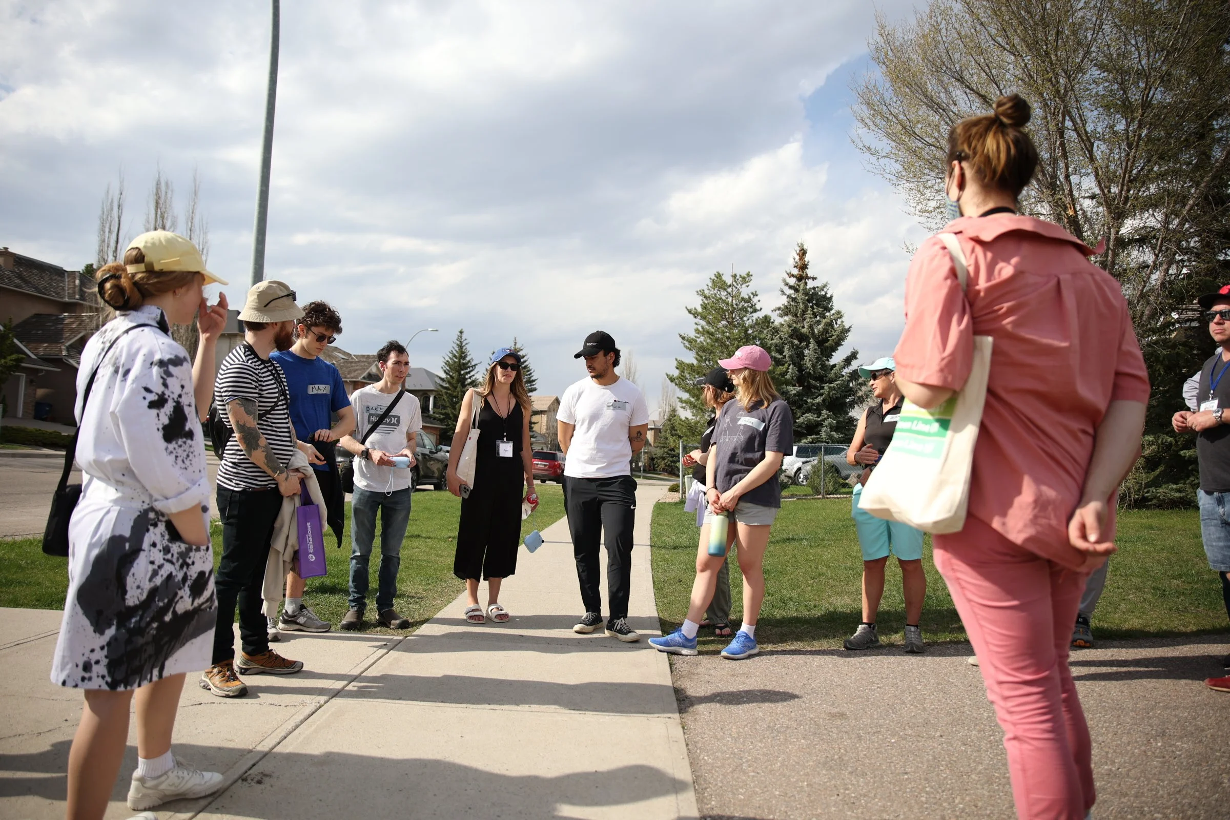 Group of people standing outdoors on a sidewalk, engaged in conversation, with trees, houses, and cloudy sky in the background.
