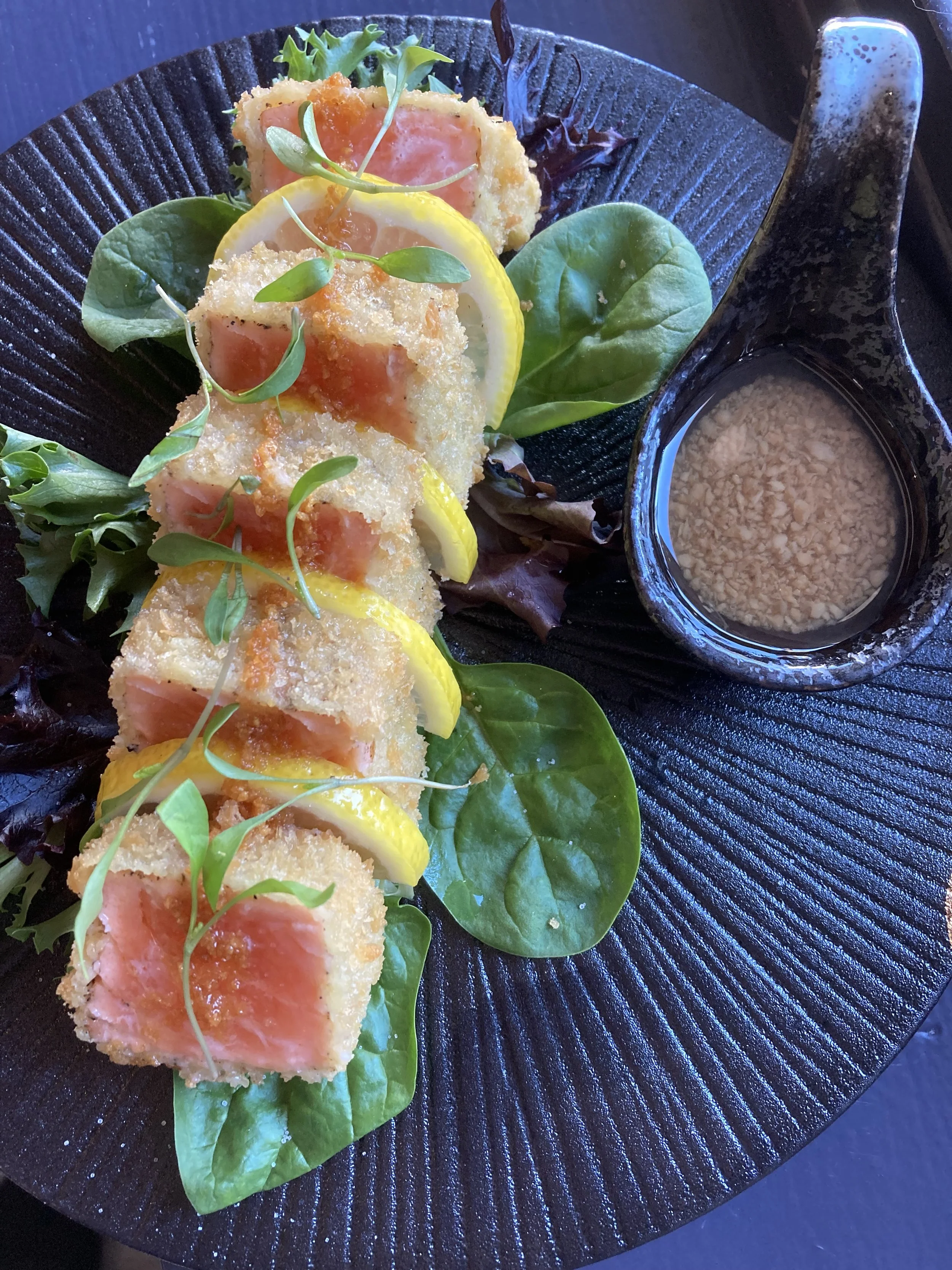 Salmon Katsu topped with lemon slices and microgreens, served with mixed greens and dipping sauce on a black textured plate.