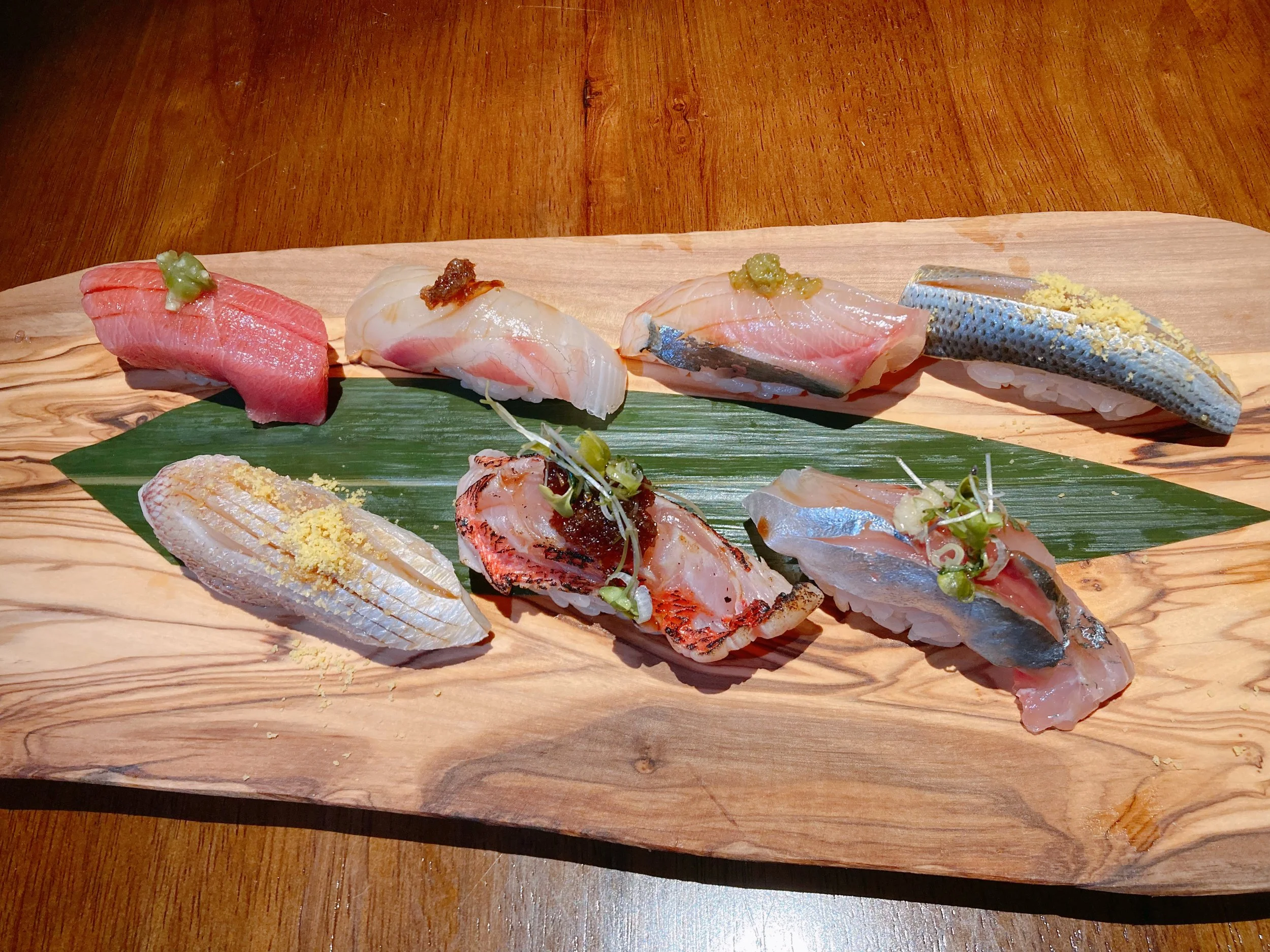 Assorted pieces of sushi with different types of fish, garnished with wasabi, on a wooden platter with a green leaf.
