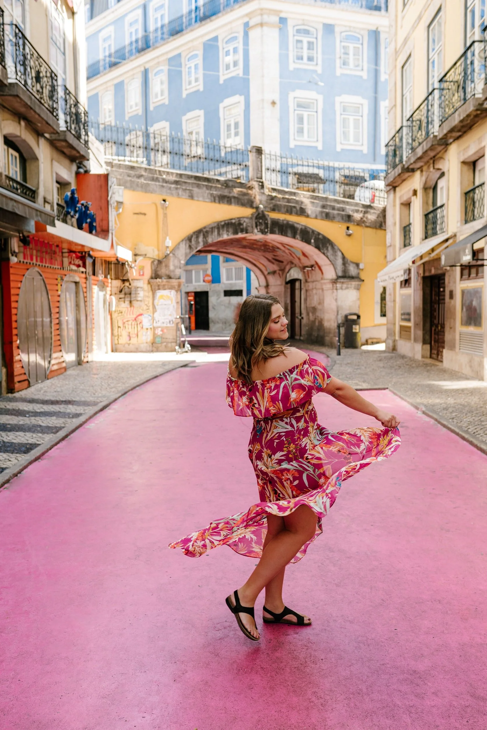 Mulher com vestido florido em movimento na rua rosa, arquitetura colorida ao fundo.