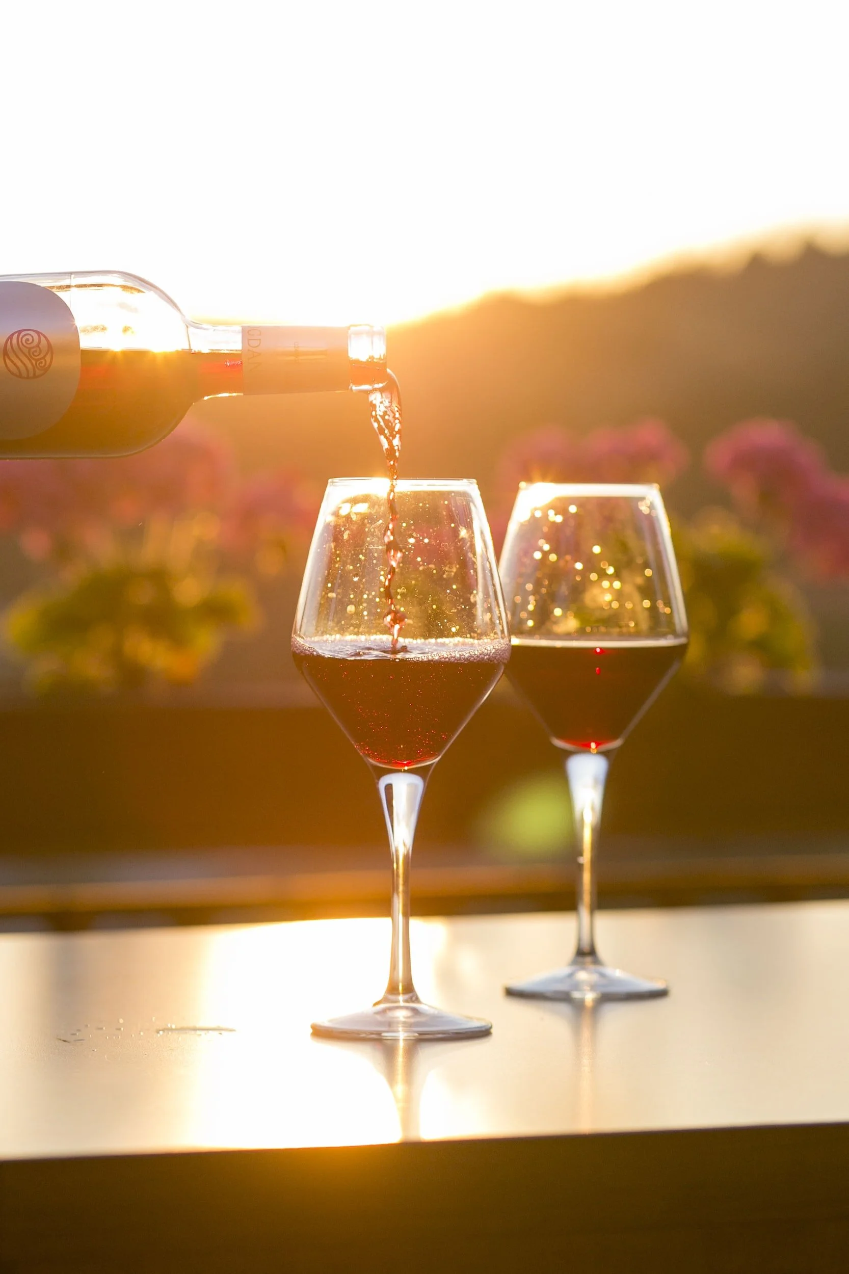 duas taças com garrafa de vinho tinto e o por do sol ao fundo