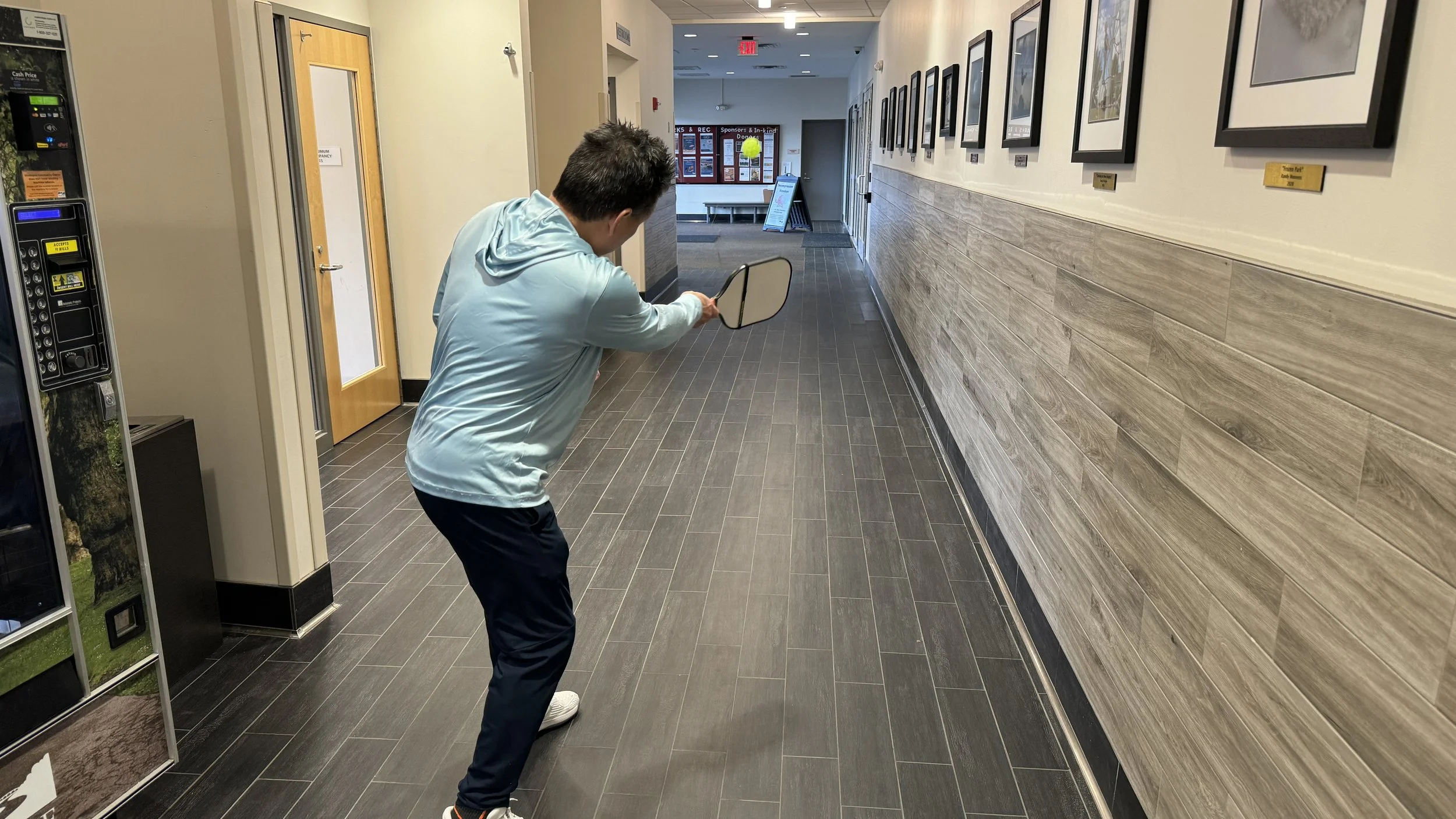 A person wearing a light blue hoodie and dark pants is hitting a mirror with a golf club in a hallway decorated with framed pictures.