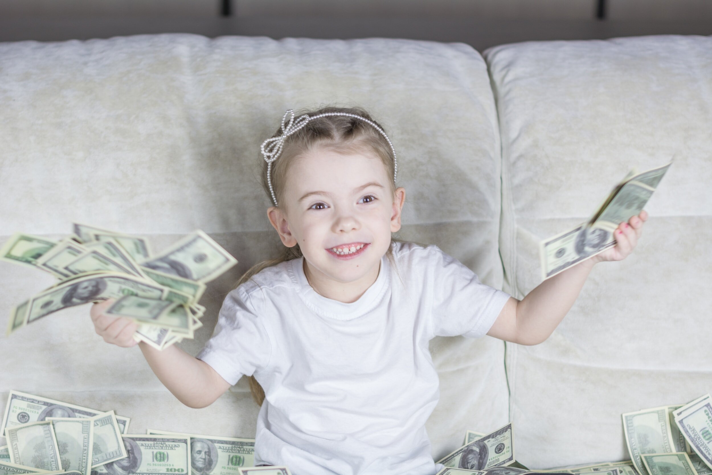 Smiling young girl with a hairband playing with lots of hundred-dollar bills on a beige sofa.
