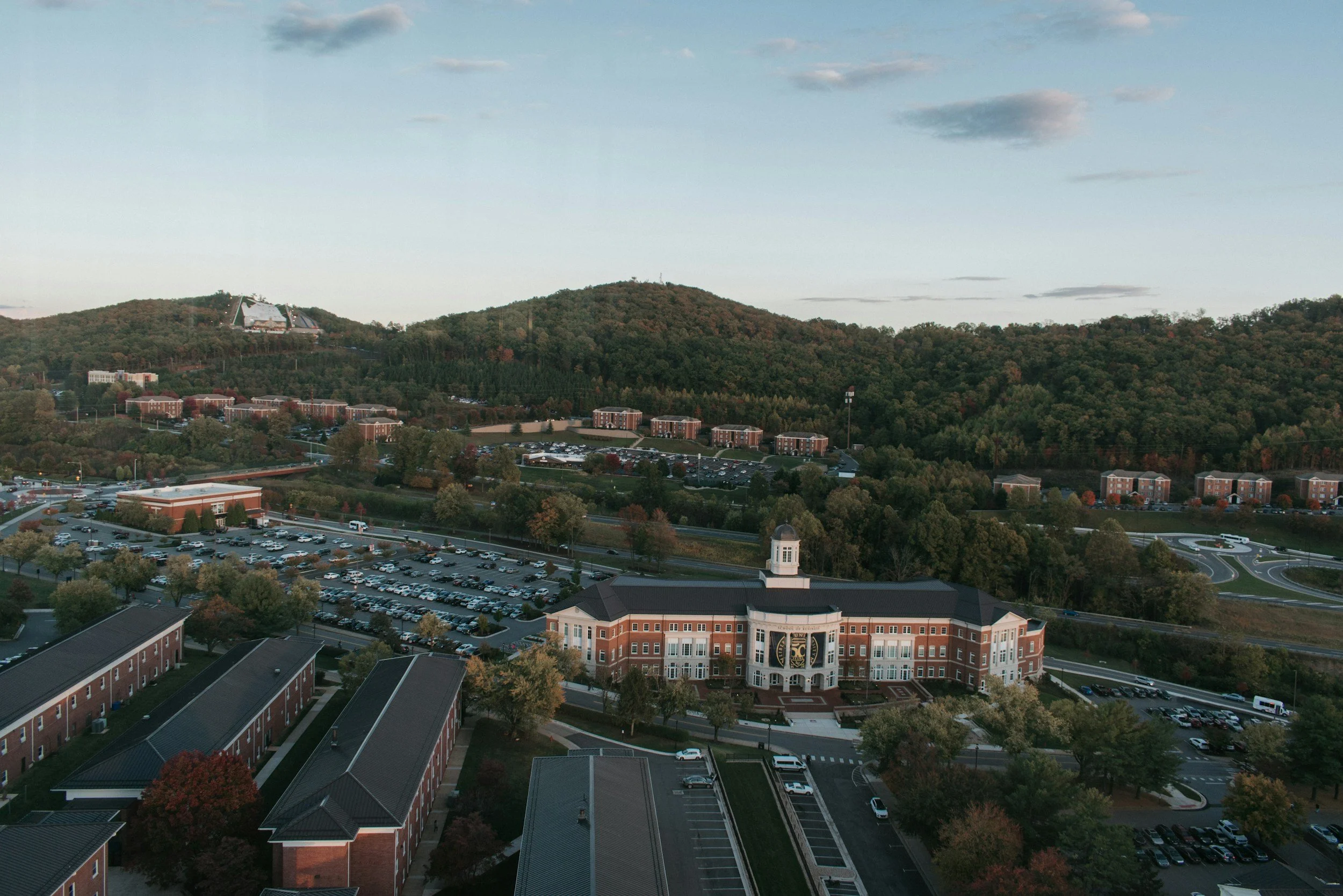 Aerial view of a university campus with brick buildings, parking lots, and surrounding trees and hills in the background during late afternoon.