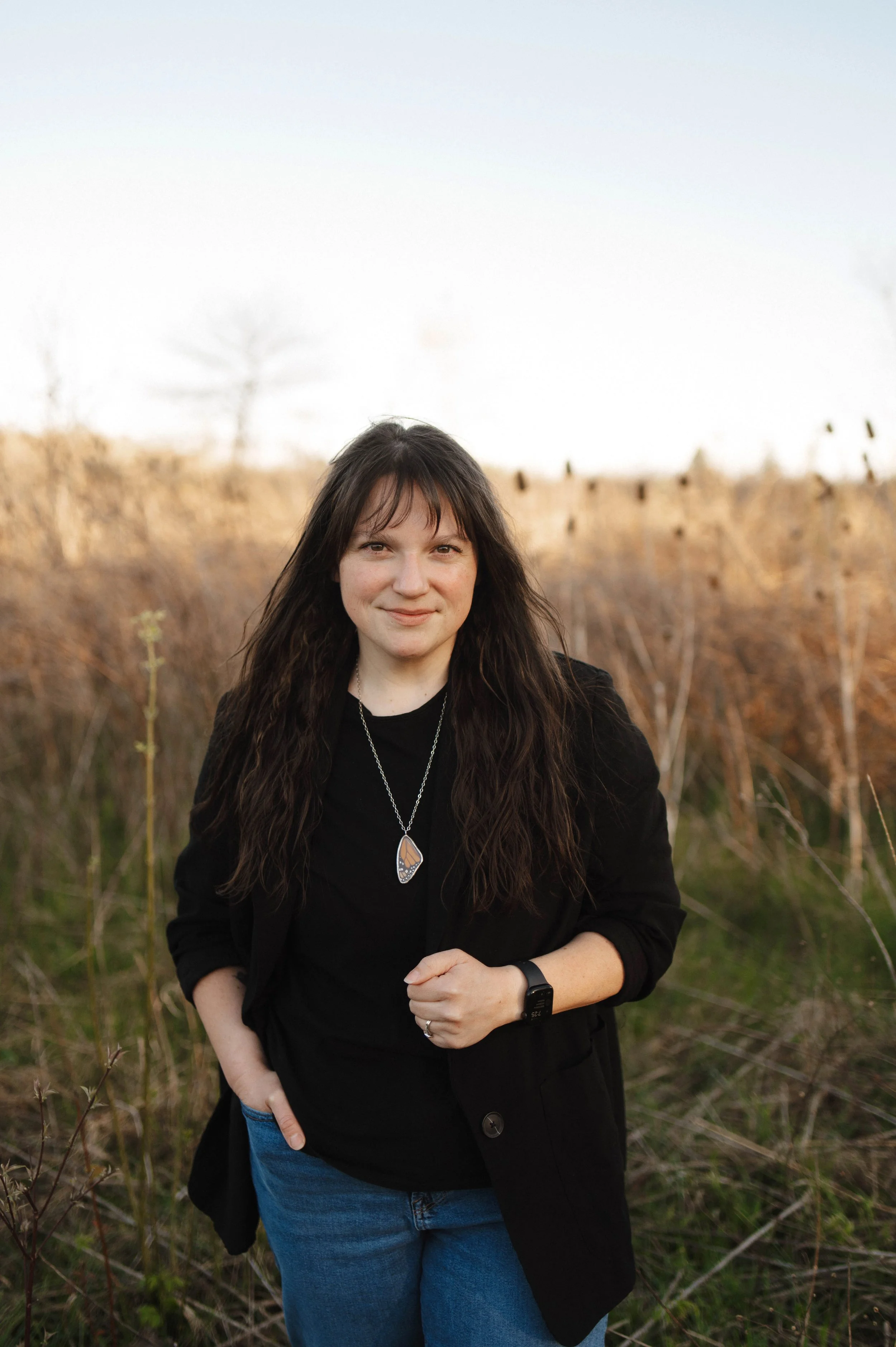 A woman with long curly dark hair standing outdoors in a field with dried tall grass, wearing a black jacket, black shirt, blue jeans, a necklace with a pendant, and a smartwatch, smiling at the camera during sunset.