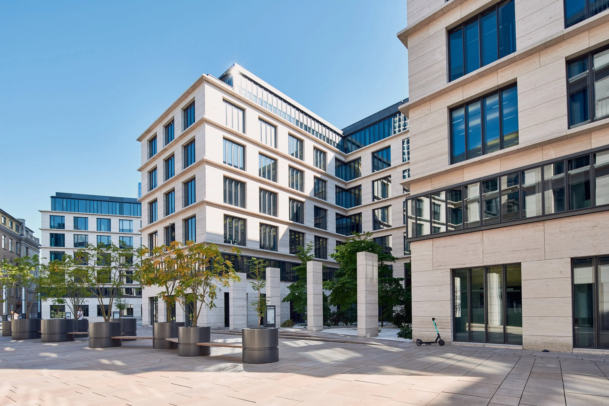 Modern multi-story buildings with large glass windows, a sunny clear sky, trees, and a scooter in a paved courtyard.