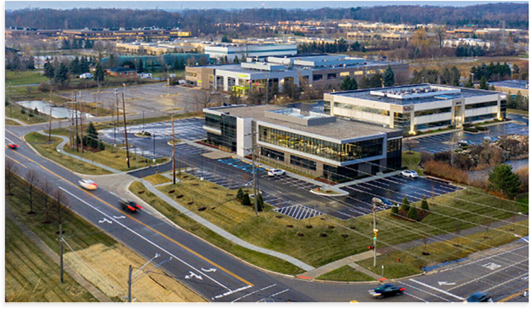 Aerial view of office buildings and parking lots along a road with moving cars in a landscaped suburban area.
