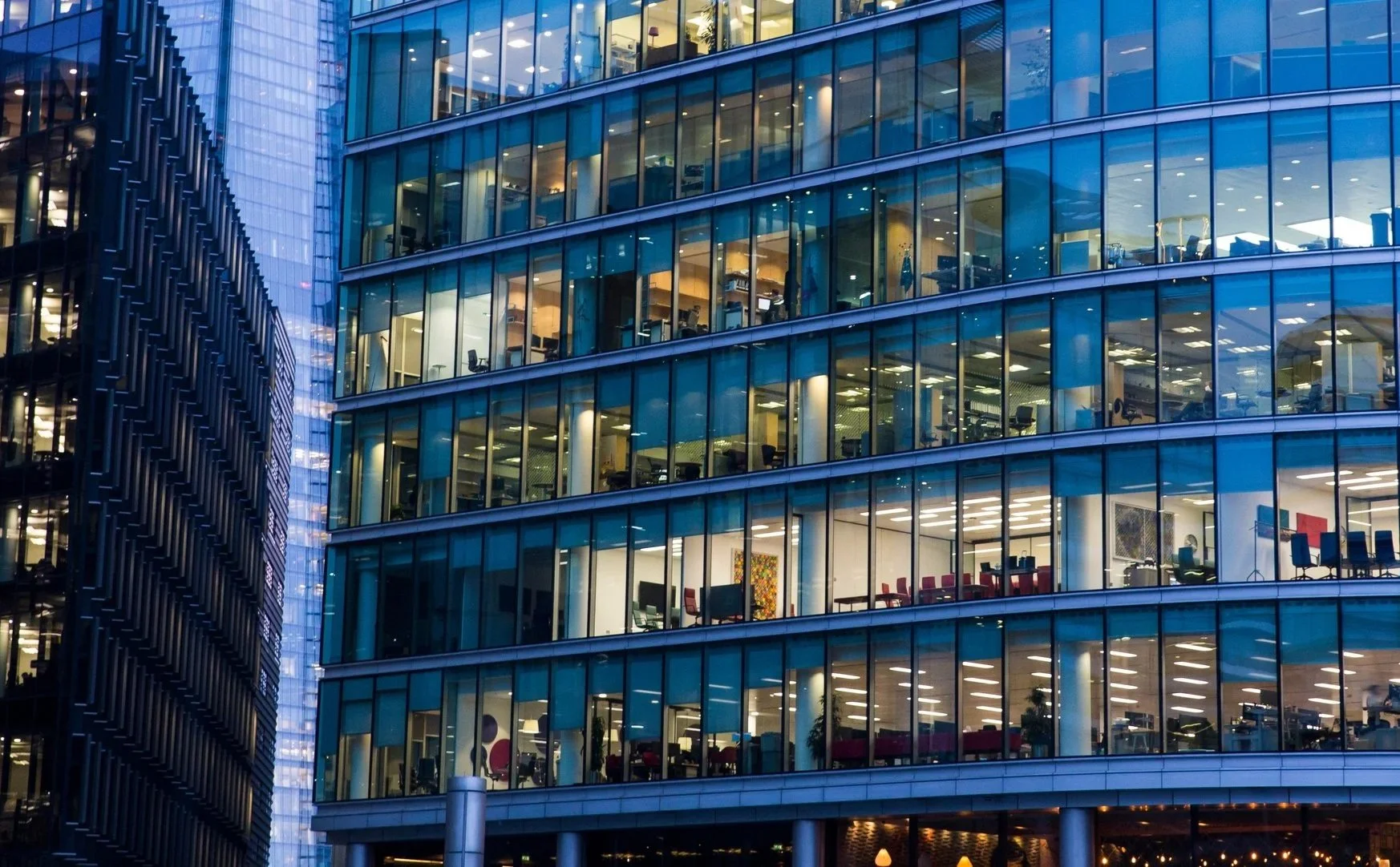 Night view of a modern glass building with illuminated office spaces visible through the windows, surrounded by other tall buildings in a city.