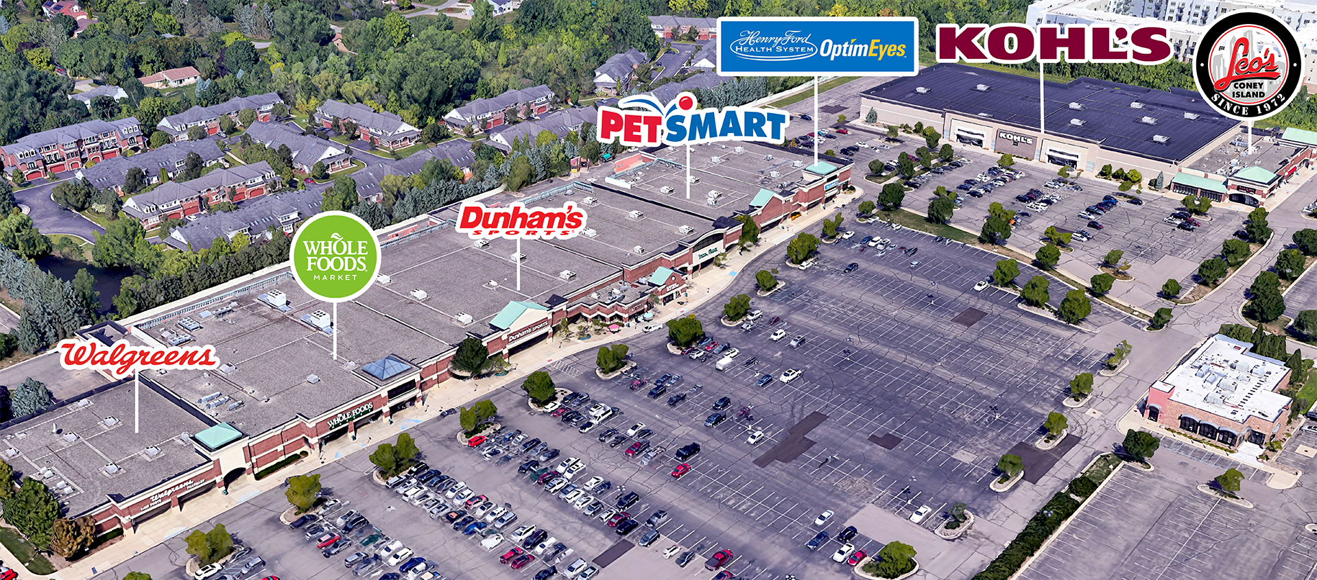 Aerial view of a shopping center with stores including Whole Foods, Walgreens, Dunham's Sports, PetSmart, Kohl's, and Leo's Coney Island. The parking lot is partially filled with cars and the area is surrounded by greenery and residential buildings.