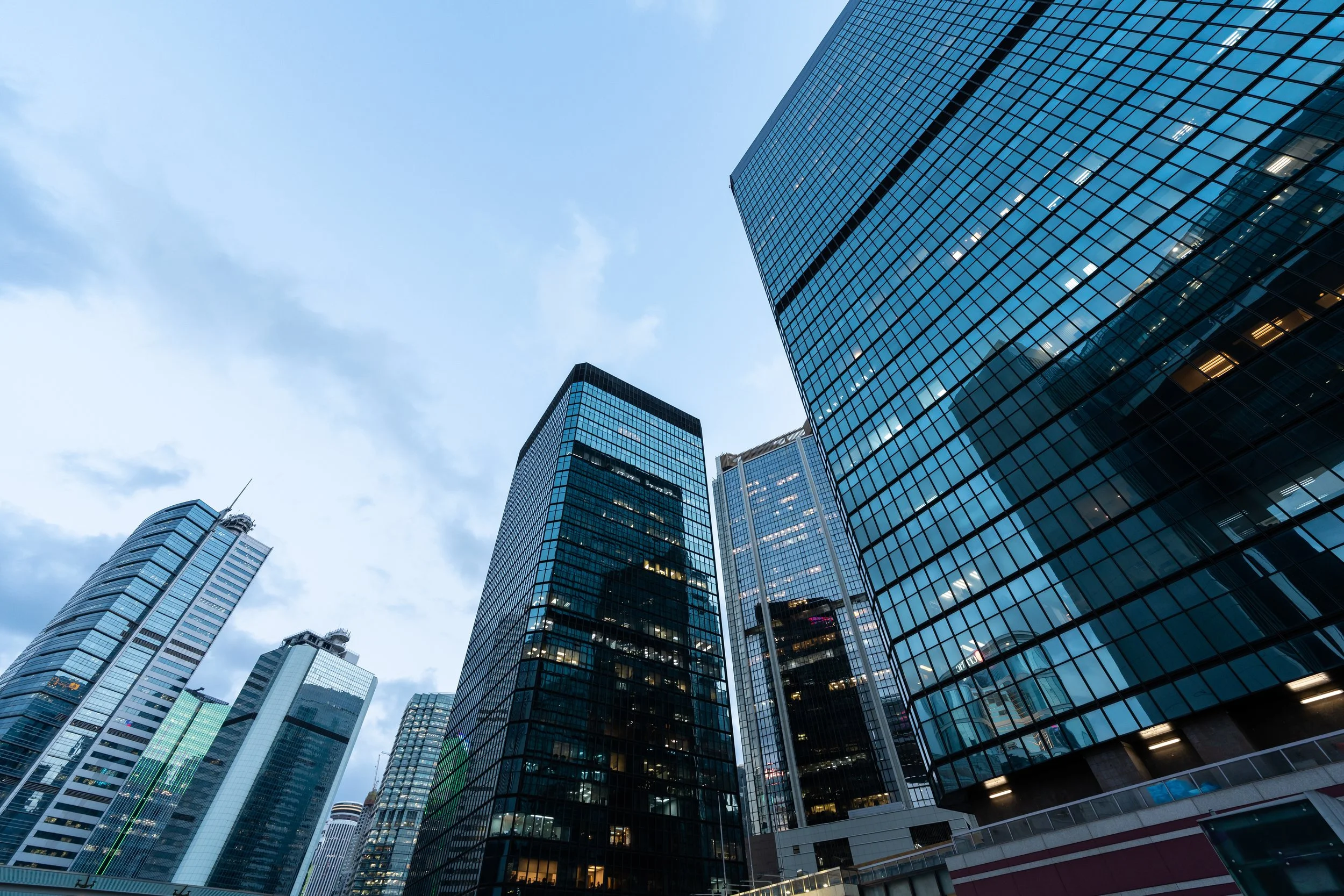 A view of tall glass skyscrapers in a city, taken from ground level looking up at the buildings against a cloudy sky.