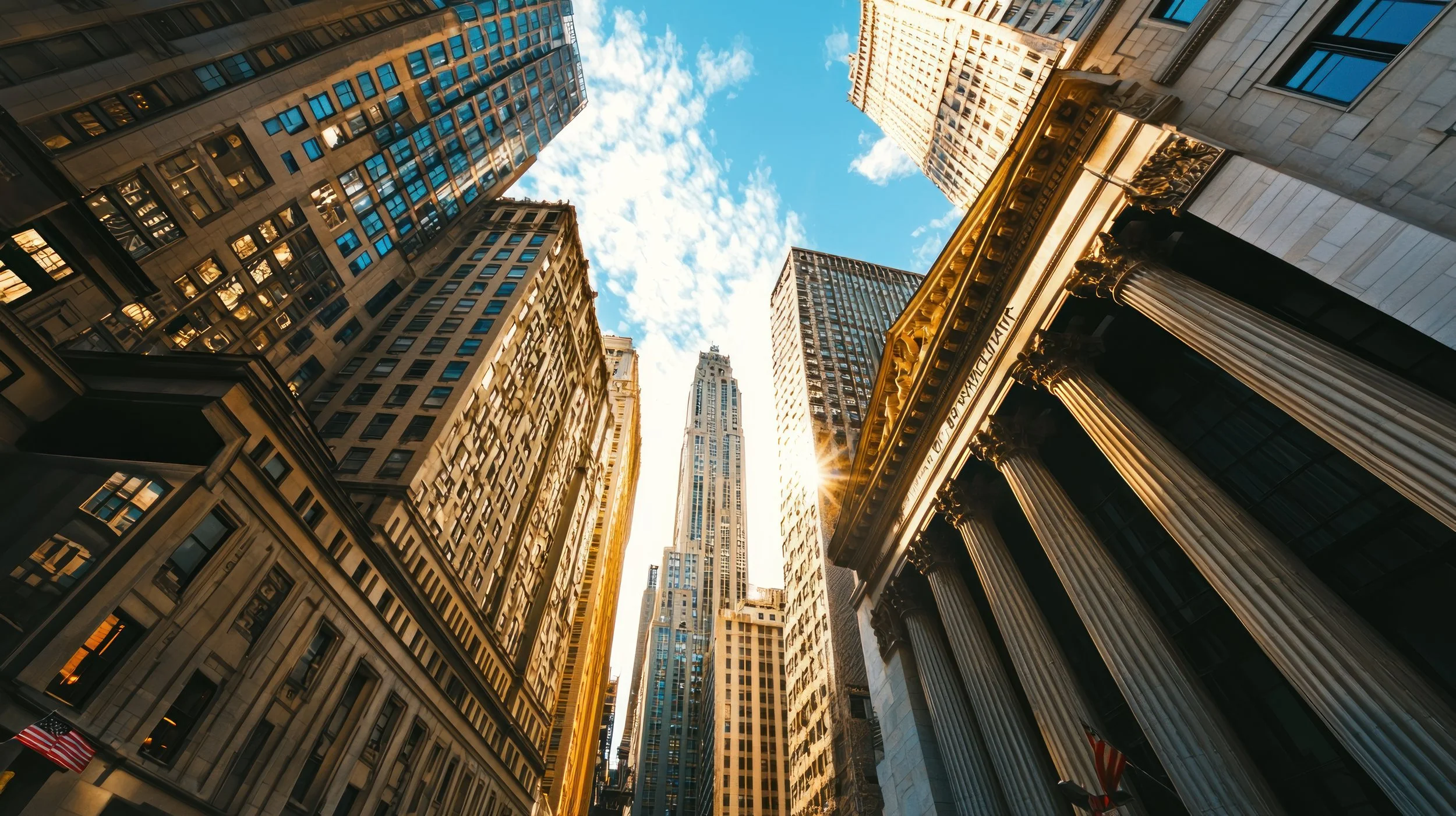 Looking up at tall skyscrapers in a city with a bright blue sky and some clouds overhead, sunlight reflecting off the buildings.