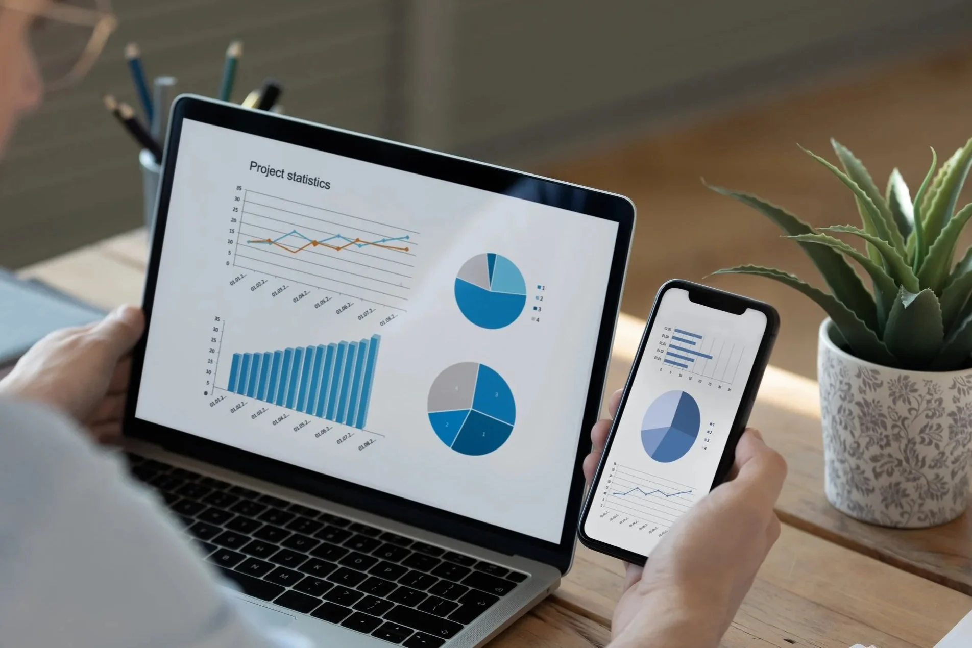 Person holding a smartphone displaying charts and graphs next to a laptop showing project statistics and data visualizations.