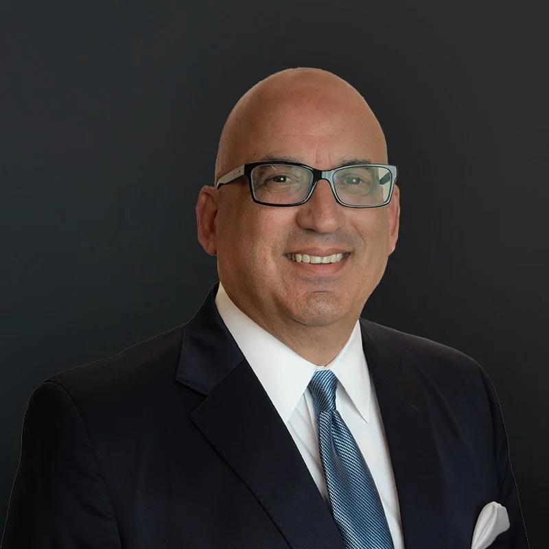 Man wearing glasses and a suit with a blue tie, smiling against a dark background.