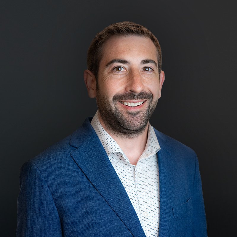 A man in a blue suit and white shirt smiling against a black background.