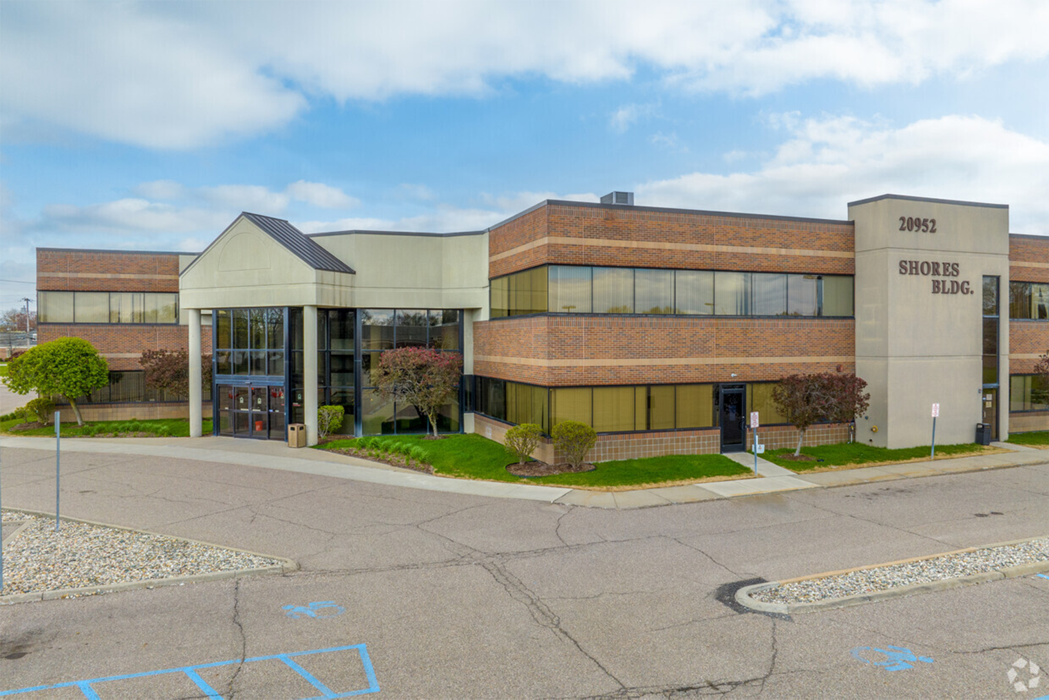 Exterior view of a two-story commercial office building labeled 'Shores Bldg.' with brick and glass facade, surrounded by landscaped greenery and parking lot.