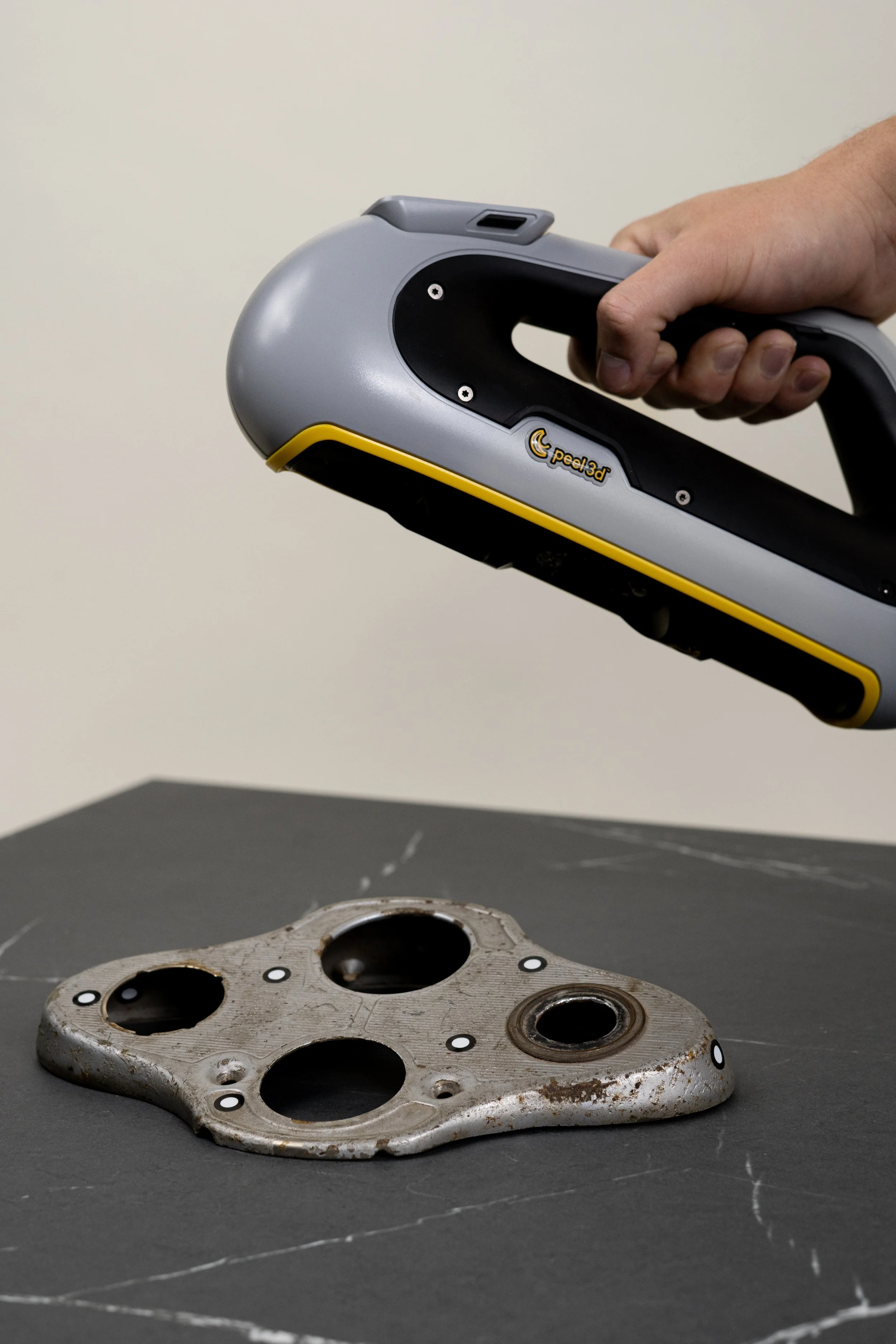 A person using a handheld 3D scanner to scan a metal motorcycle part with multiple holes and rust spots, placed on a black surface.