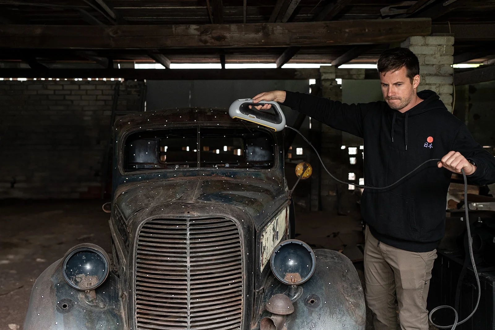 Man using a handheld scanner or inspection device on an old, rusty vintage car inside a garage or workshop.