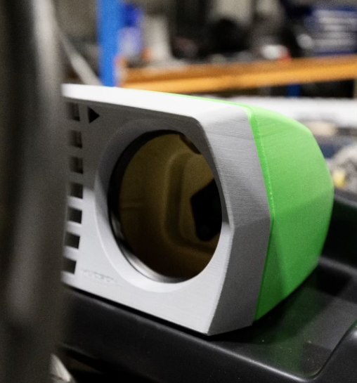 A device with a white front, a large circular opening, and green side panel, placed on a black surface in a workshop or garage setting.