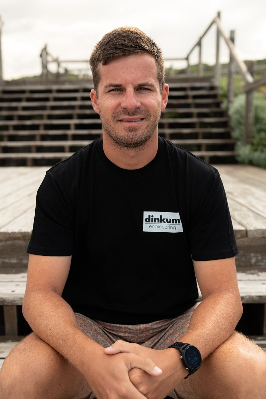 A man wearing a black T-shirt with the text 'dinkum engineering' sits outdoors on wooden steps, smiling at the camera.