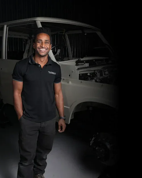 Man smiling standing in front of a partially disassembled vehicle in a workshop.