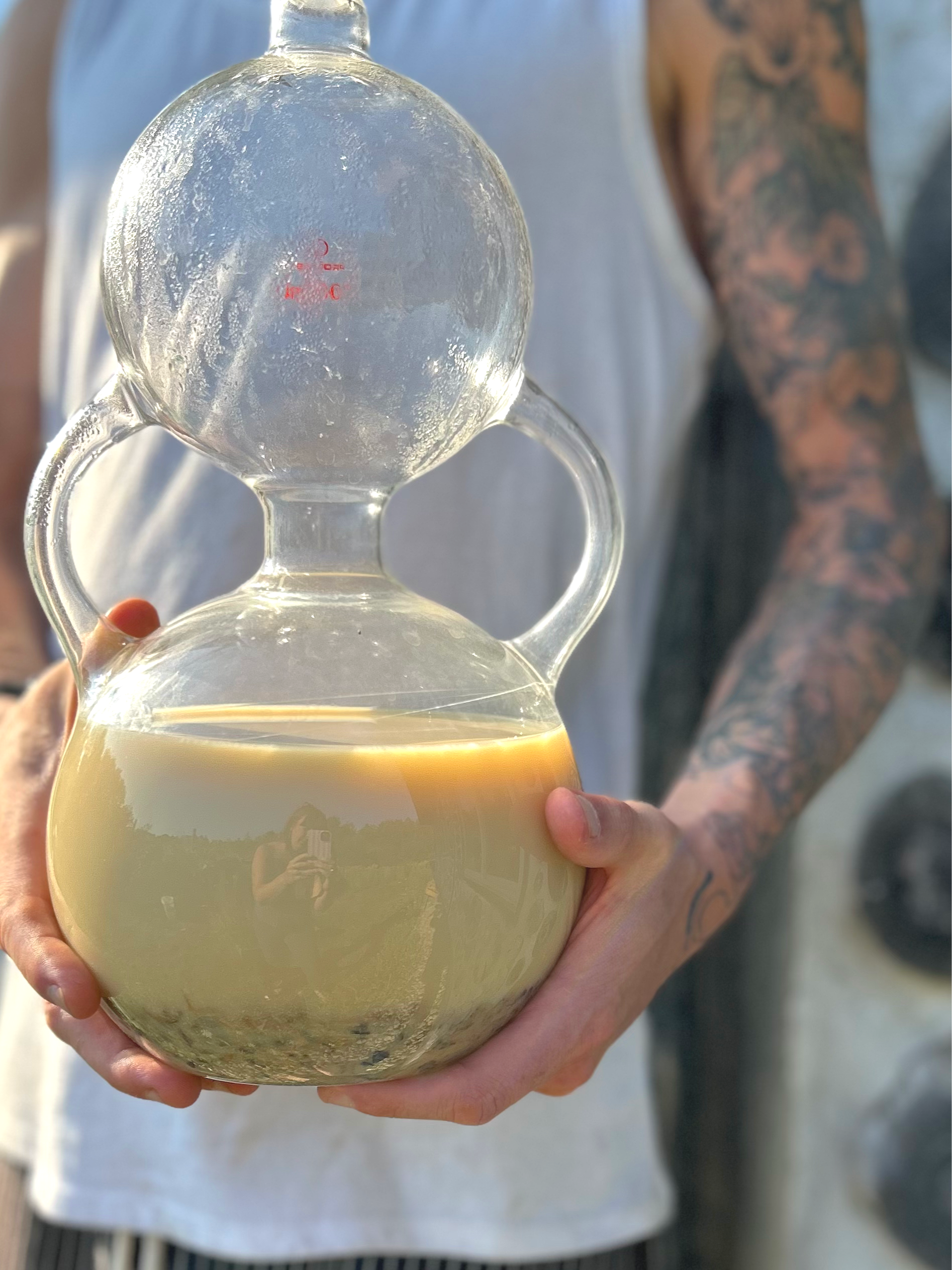 Person holding a glass teapot with a light-colored liquid inside, and a round glass top on a sunny outdoor setting. The person's arm has a tattoo, and they are wearing a sleeveless shirt.