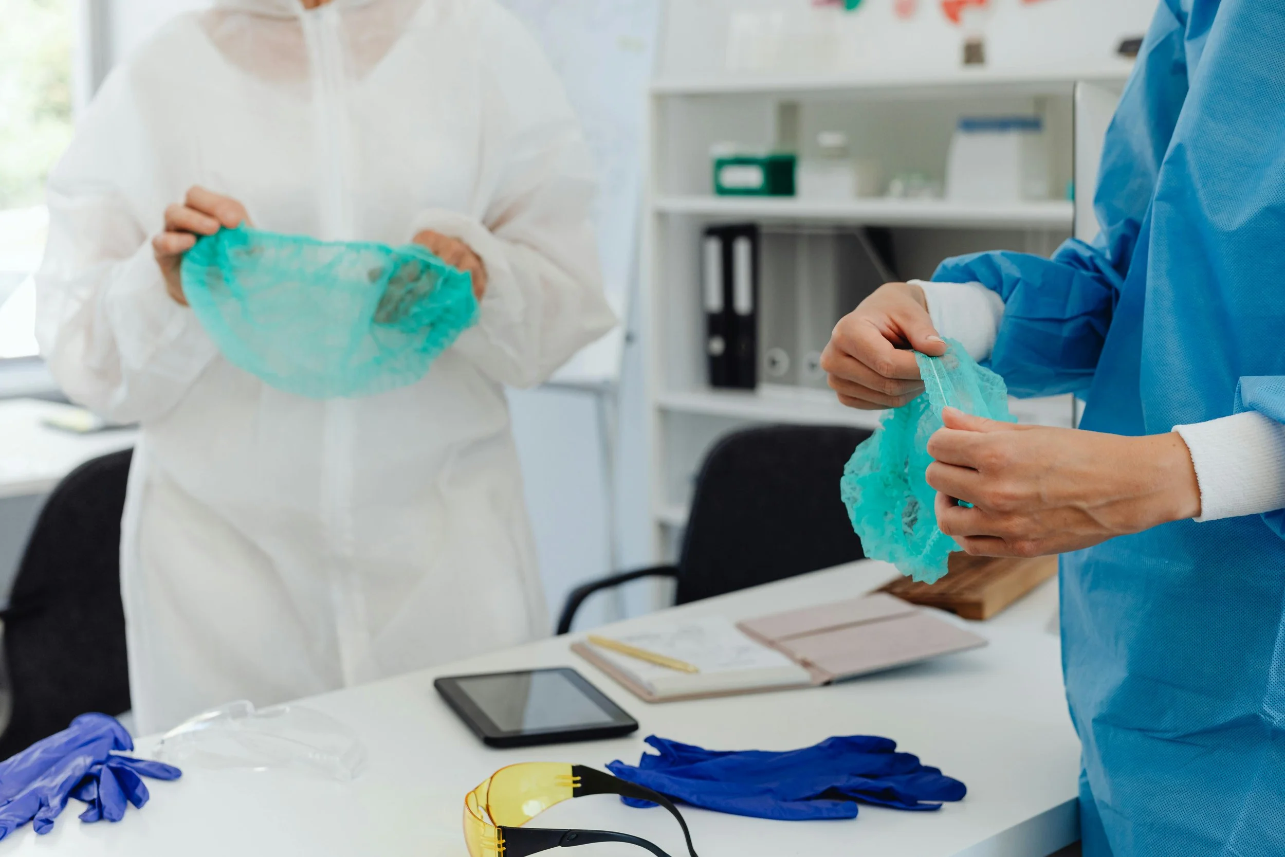 Two people in protective clothing handling medical or laboratory gloves in a clinical setting, with various equipment and tools on the table.