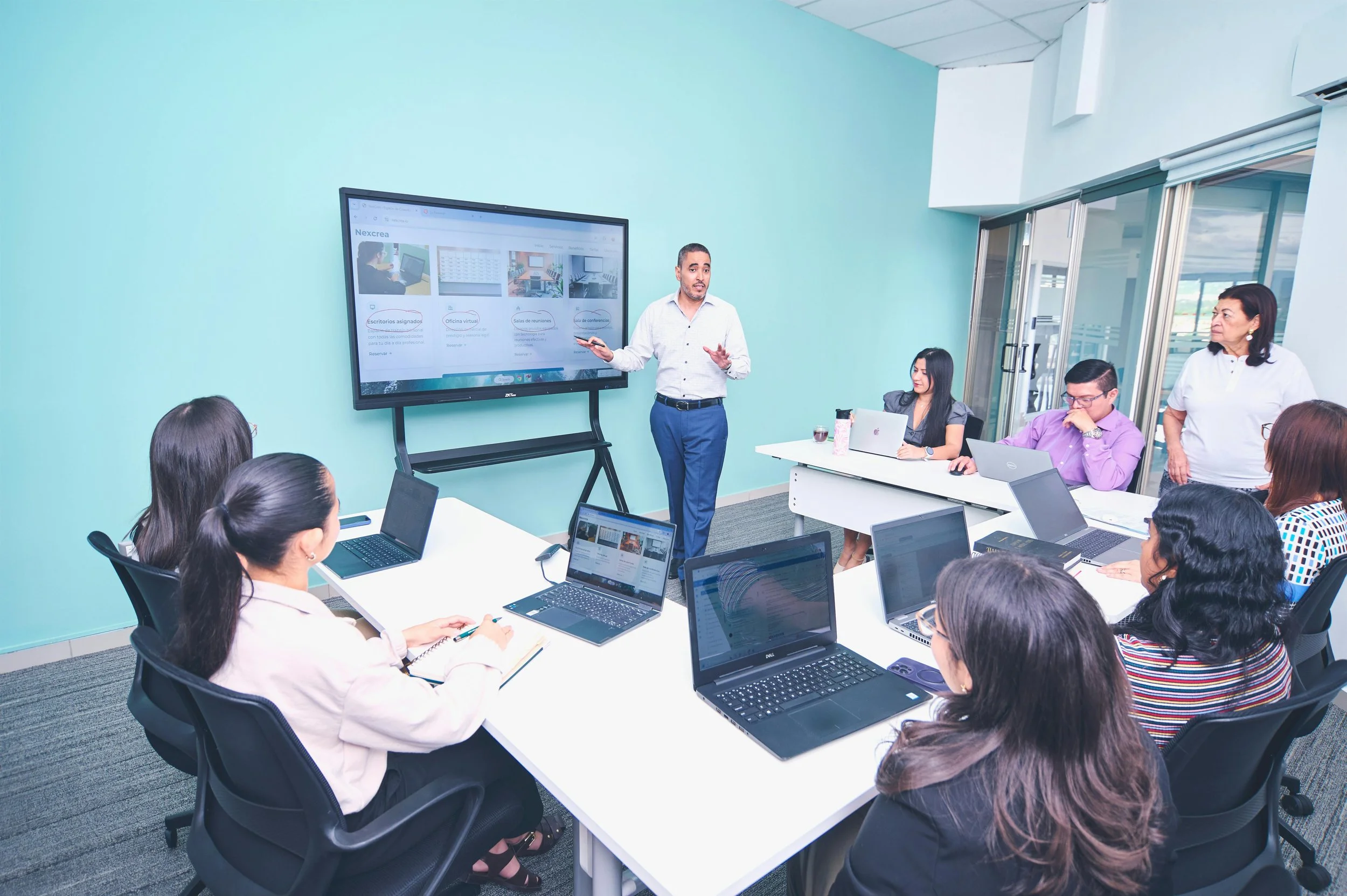 A man is giving a presentation to a group of people in a conference room. The group is seated at a U-shaped table with laptops, and the presenter is standing next to a large screen displaying a website or digital content.