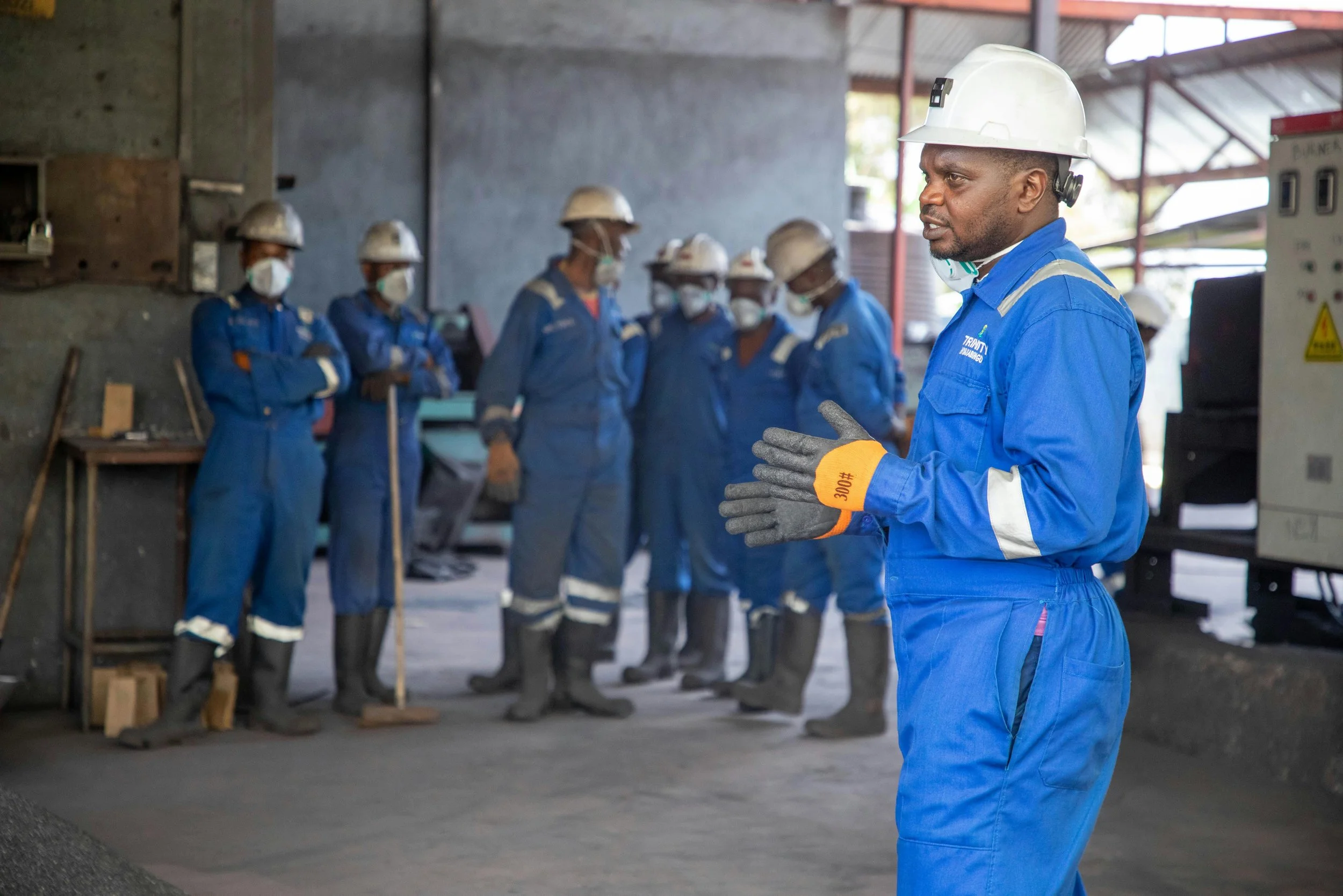 A group of construction workers wearing blue uniforms, white helmets, and face masks inside a building, with one man in the foreground giving instructions.