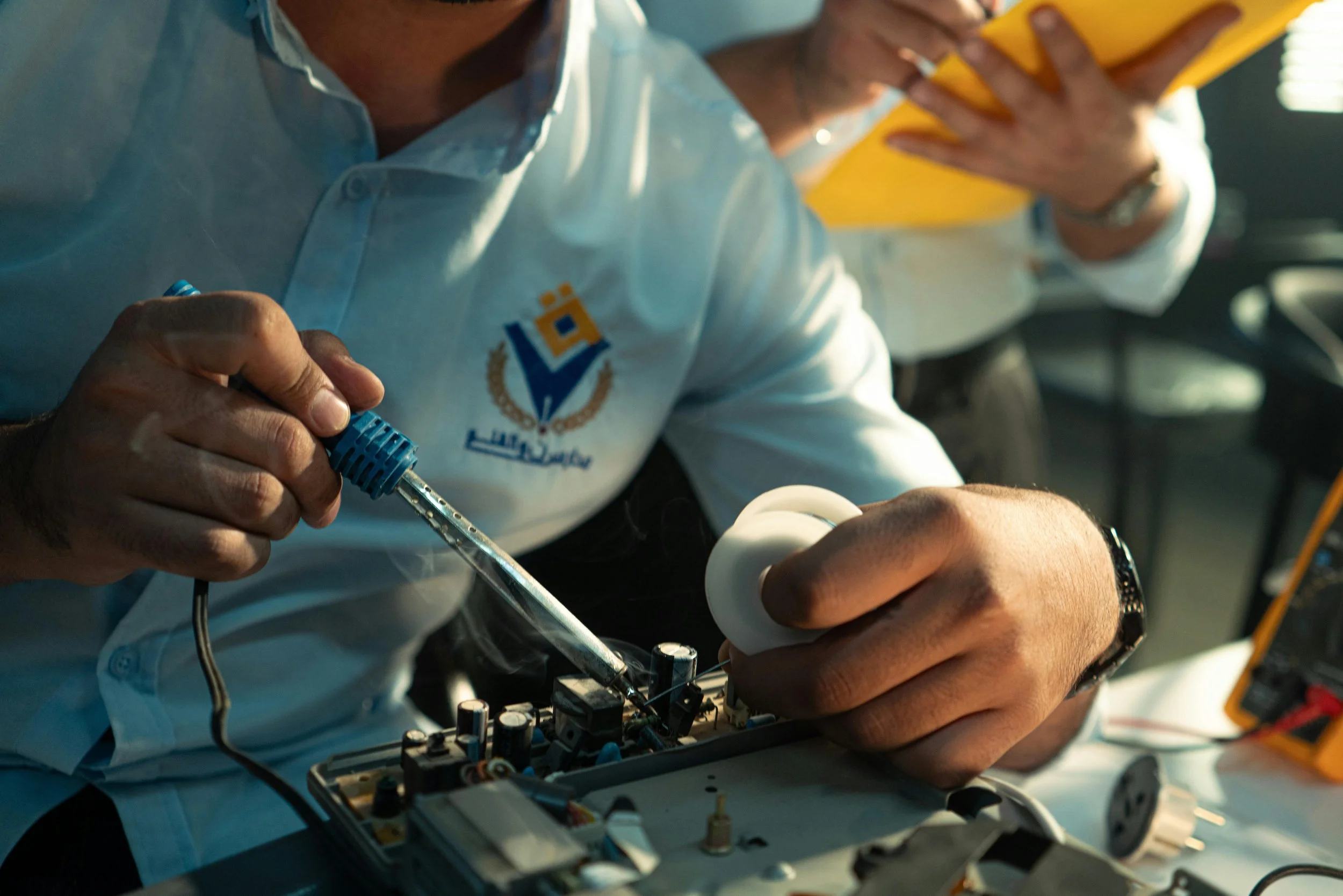 A person in a white shirt with a logo working on electronic components, possibly repairing or assembling a device