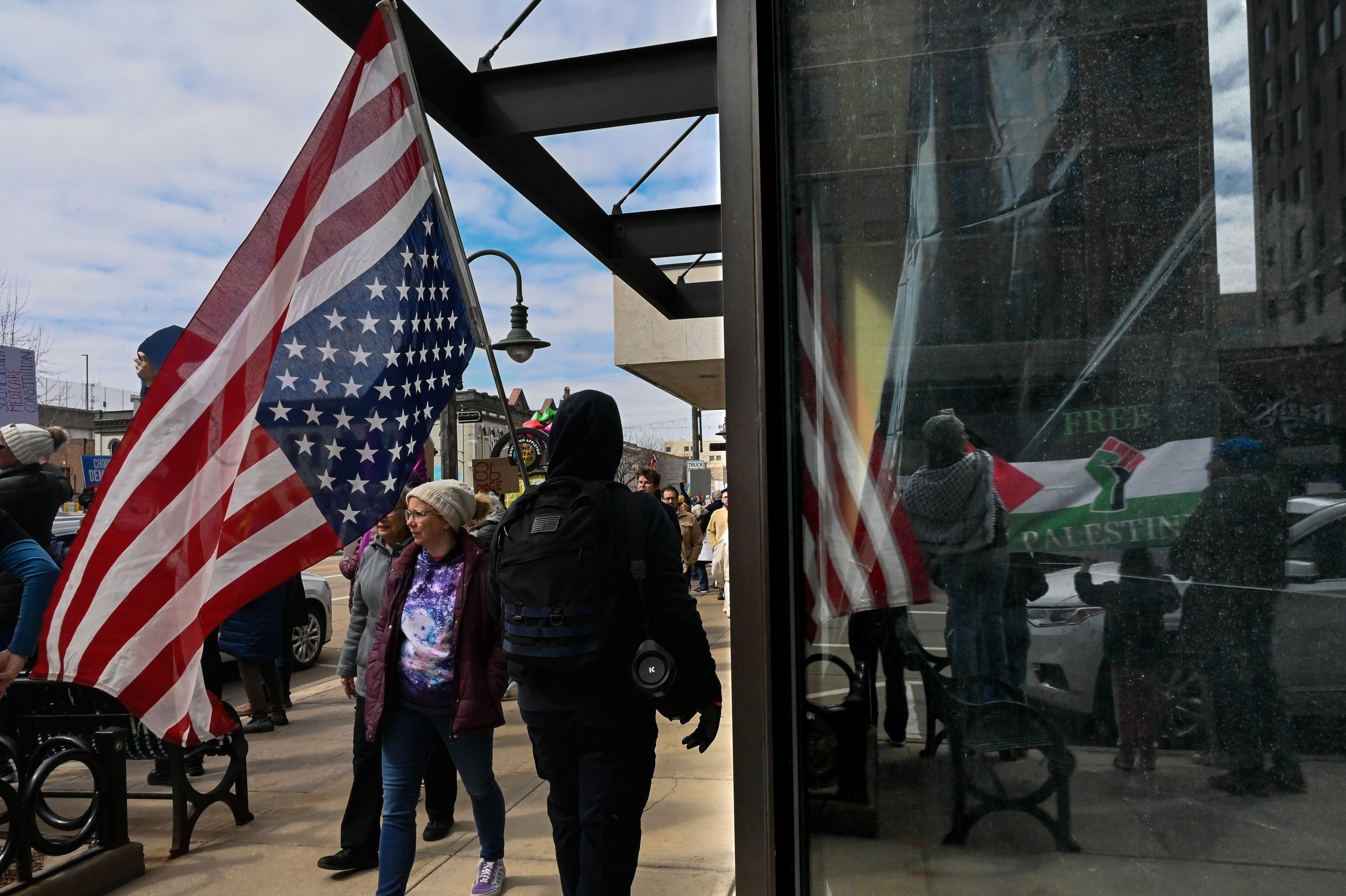 A man walks by flying an inverted American flag as a family holding a Palestinian flag is reflected in the window at Houdini Plaza in Appleton, Wisconsin, on April 5, 2025. The protest was part of a mass mobilization of over 1200 protests in all 50 s