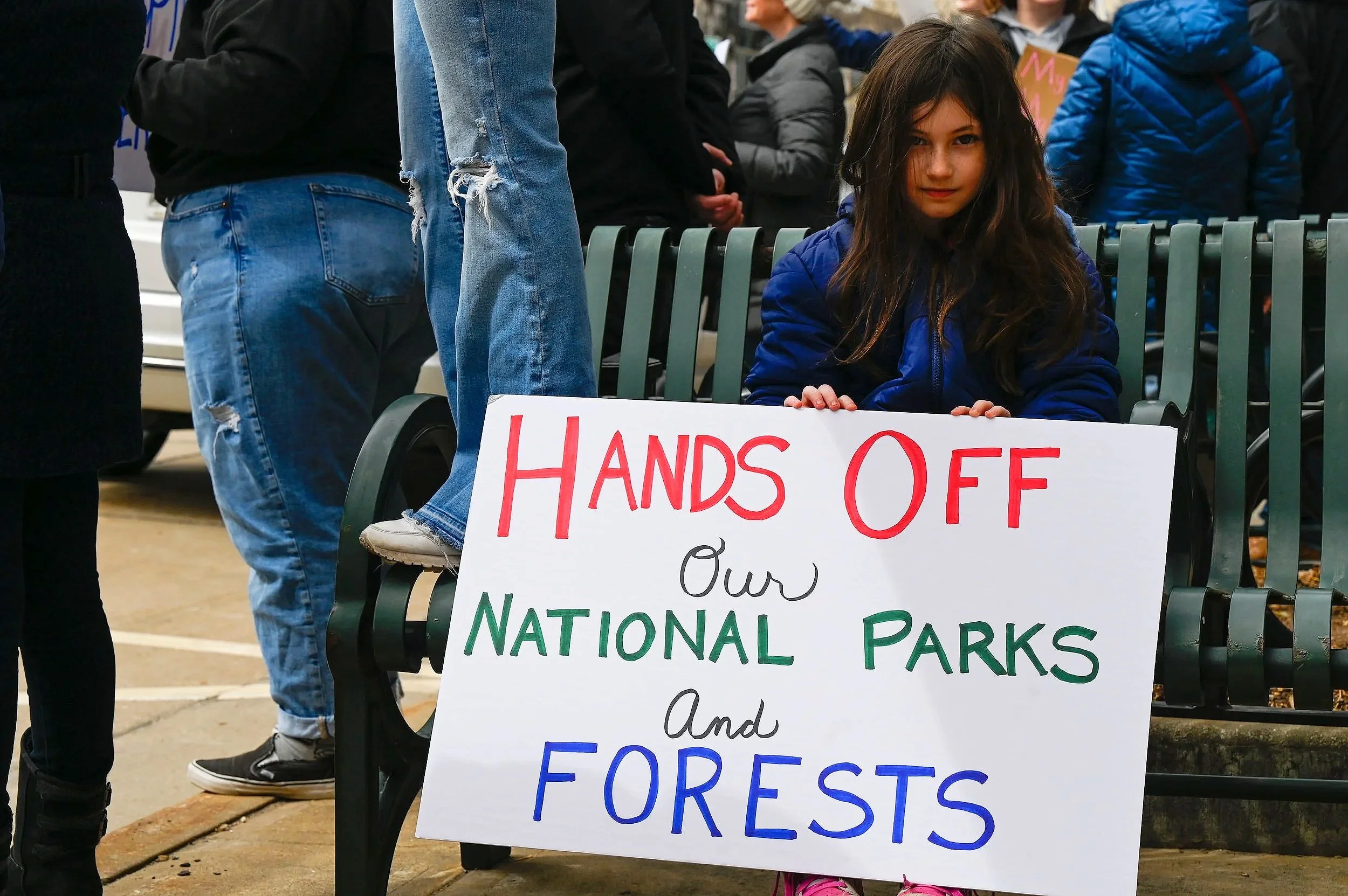 A young girl holds a sign labeled "Hands Off Our National Parks and Forests" while her family stands beside her raising a "Free Palestine" flag at Houdini Plaza in Appleton, Wis., on April 5, 2025. Around 1,000 National Park Service employees were la