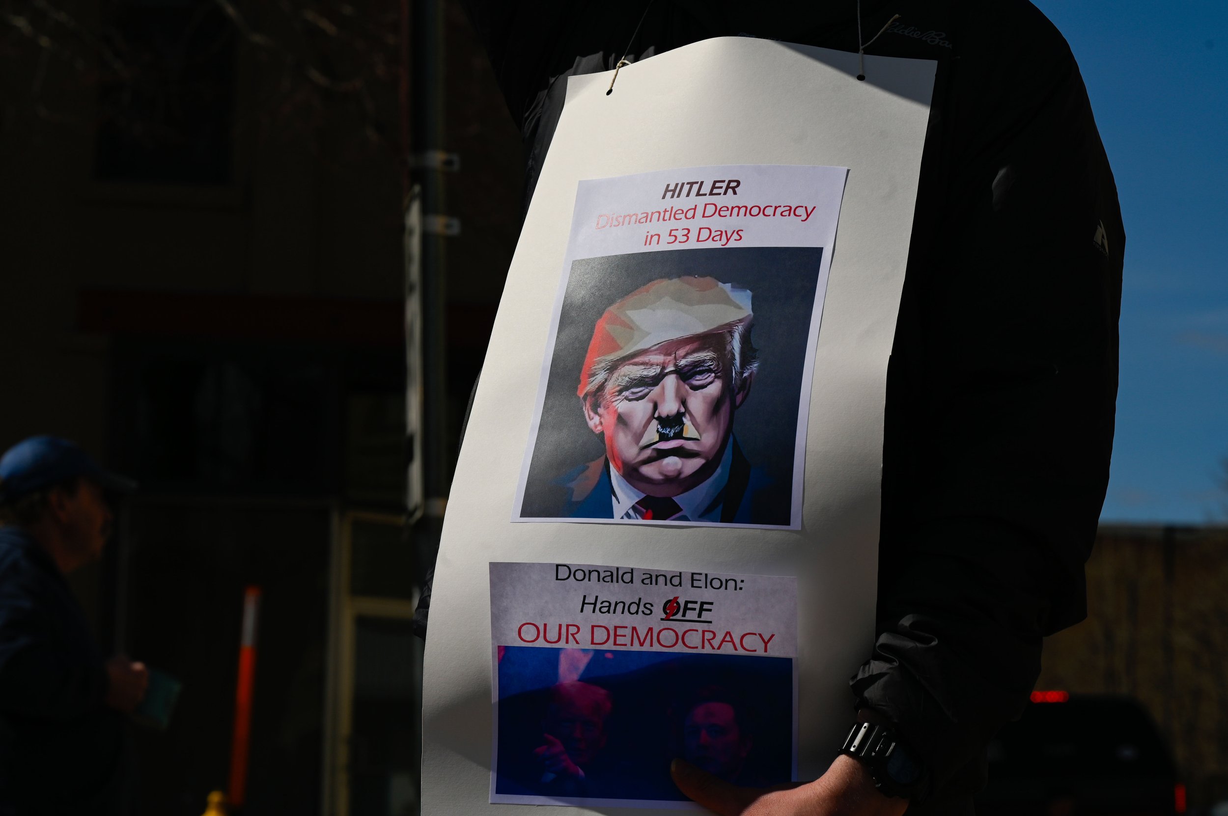 A man wears a sign tied around his neck that says "HITLER Dismantled Democracy in 53 Days" and "Donald and Elon: Hands OFF OUR DEMOCRACY" while at a protest at Opera House Square in Oshkosh, Wisconsin on April 5, 2025. State Representative Lori Palme