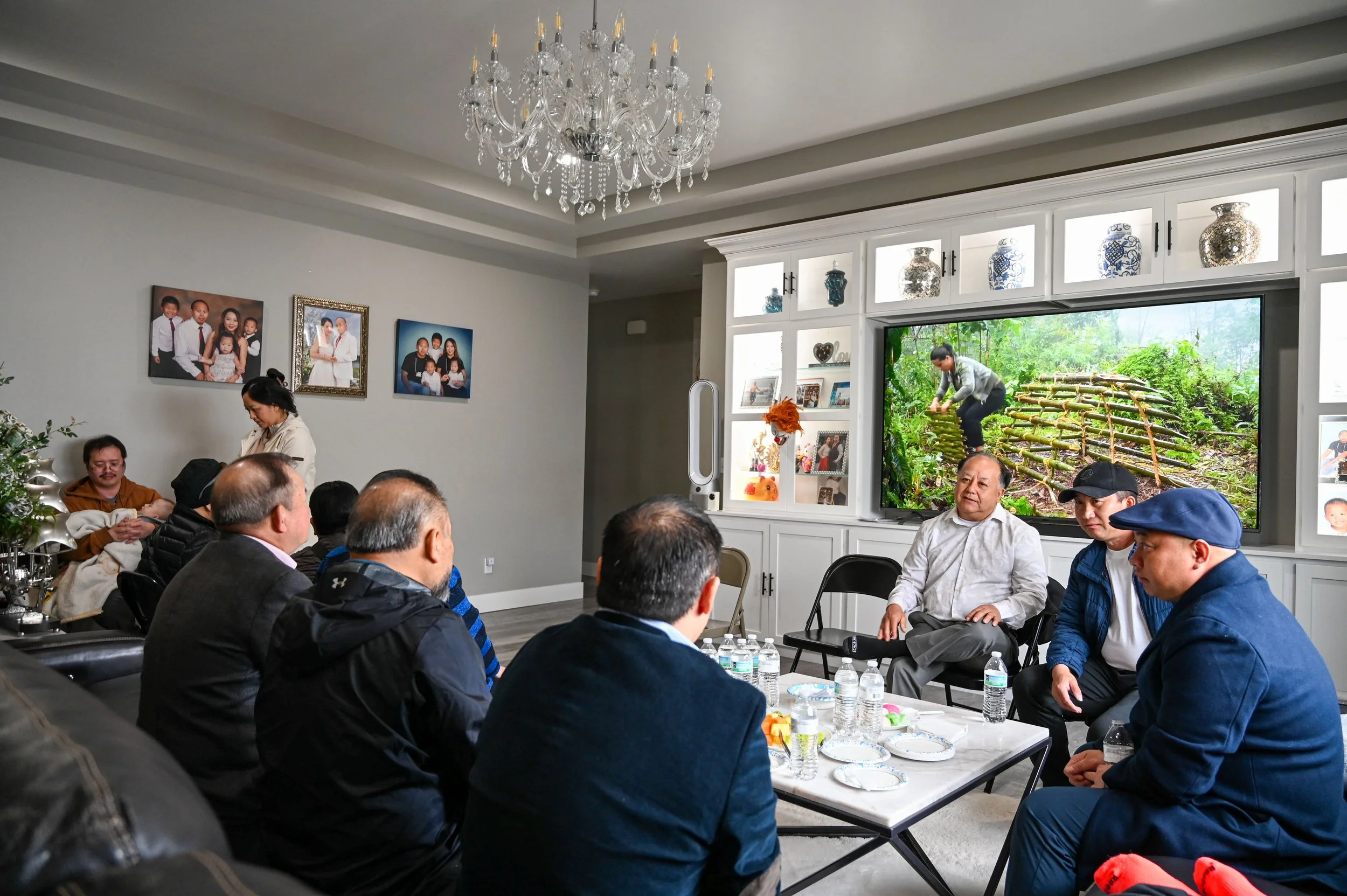 Several men gather around a table in the living room as guests arrive at an elder name-changing ceremony in La Crosse, Wis., Saturday, April 18, 2026. A younger man holds his baby as his partner stands by. In Hmong culture, it is common for women to 