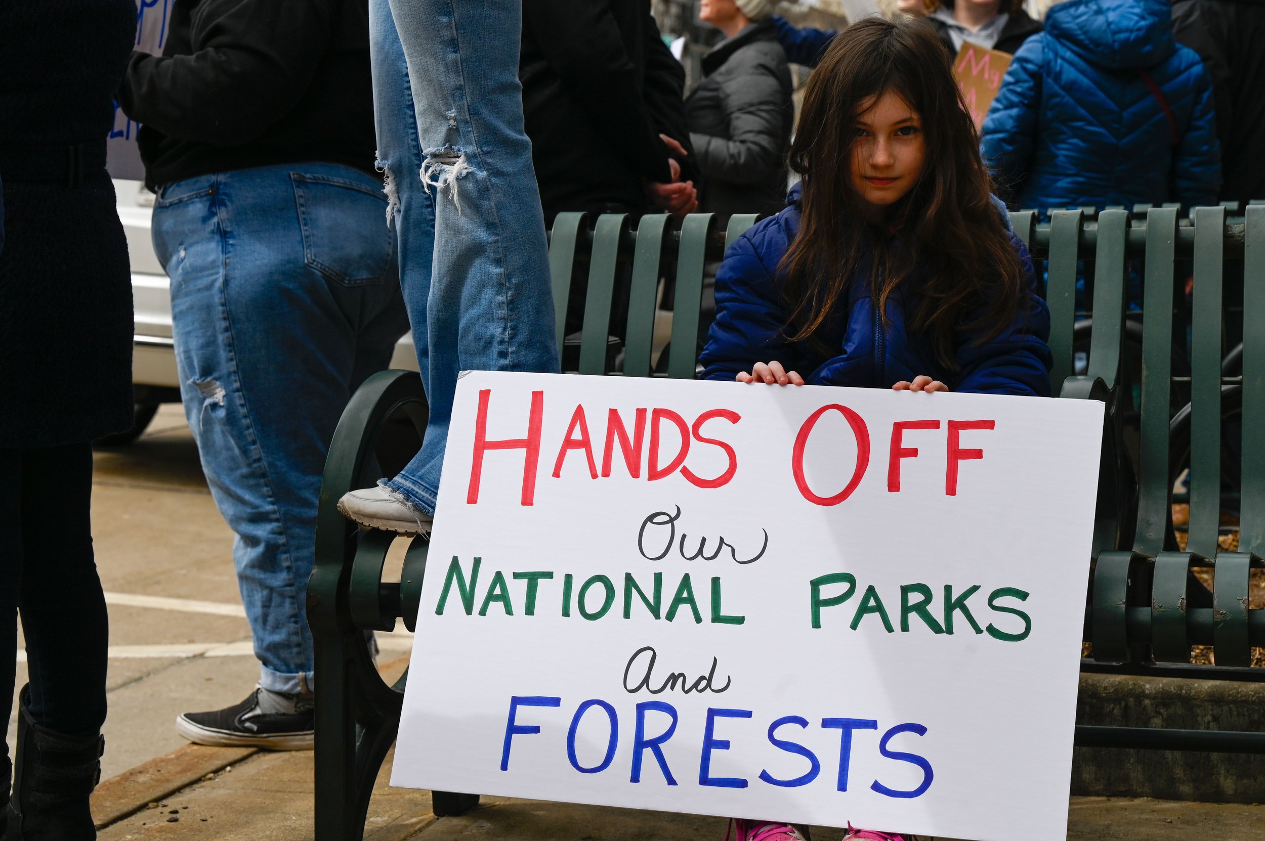 A young girl holds a sign labeled "HANDS OFF Our NATIONAL PARKS and FORESTS" while her family stands beside her raising a "Free Palestine" flag at Houdini Plaza in Appleton, Wisconsin, on April 5, 2025. Around 1,000 National Park Service employees we