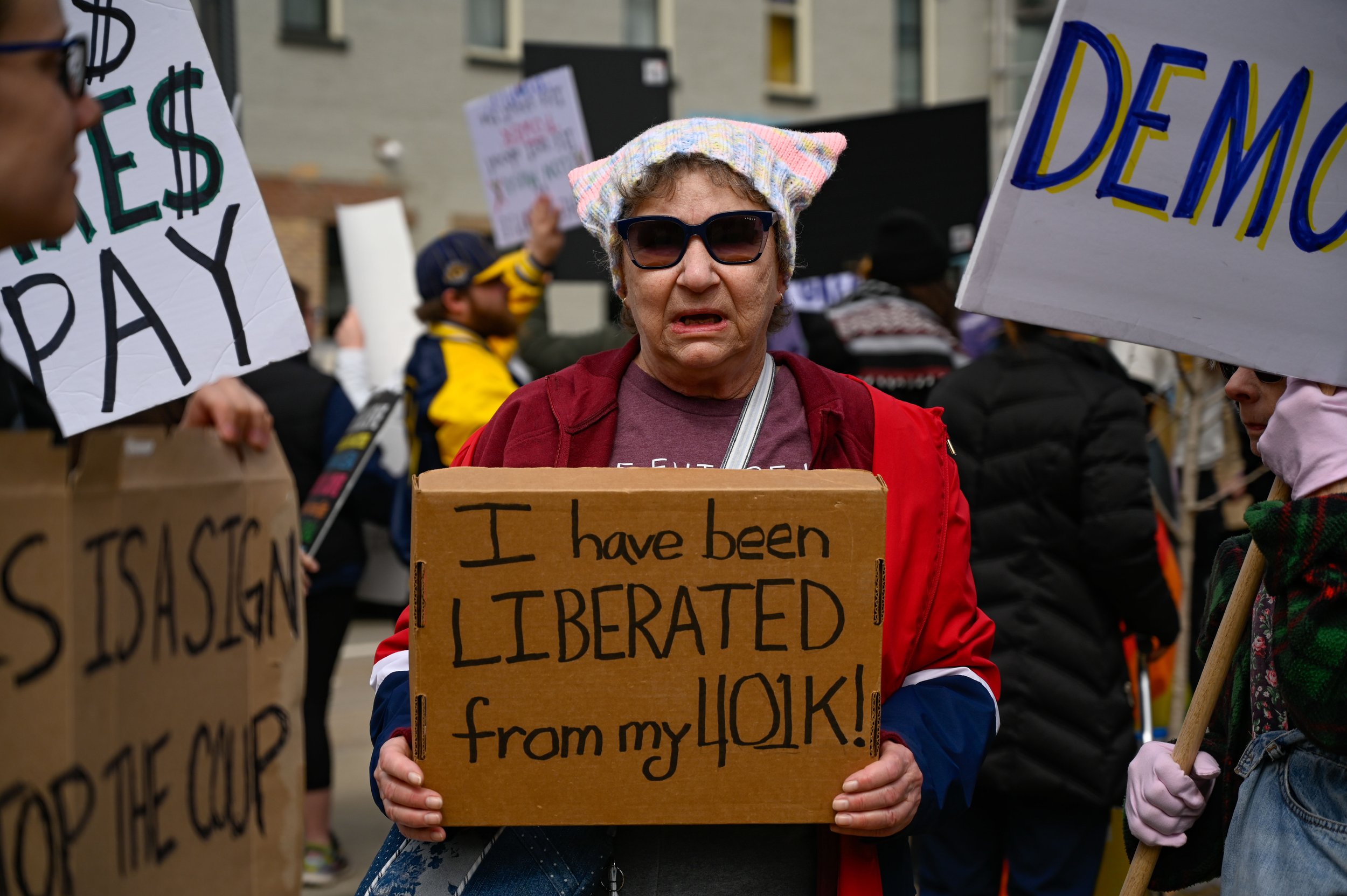 A woman wearing a "Pussyhat" holds a sign in a crowd of protesters at Houdini Plaza in Appleton, Wisconsin, on April 5, 2025. The "Hands Off!" protest was to oppose the actions of President Donald Trump and his counterpart, Elon Musk. The "Pussyhat P