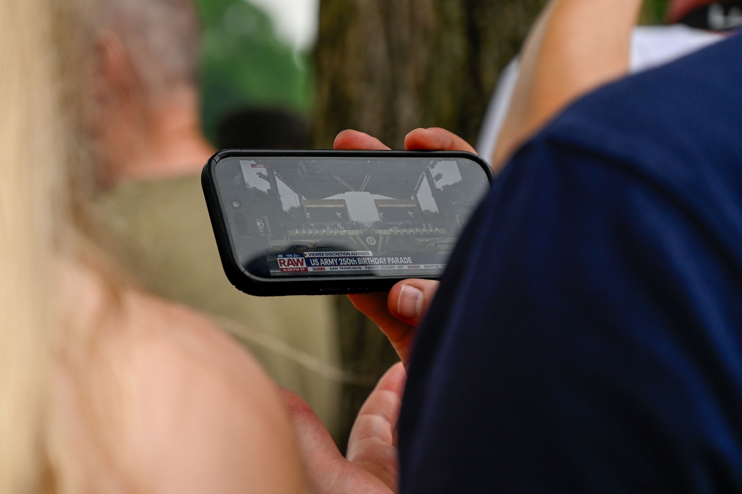 A group of attendees at the Army's 250th birthday celebration watch the opening address on a phone in Washington, D.C., on June 14, 2025. The group stood in front of a barrier down the road from the stage where President Trump was seated.