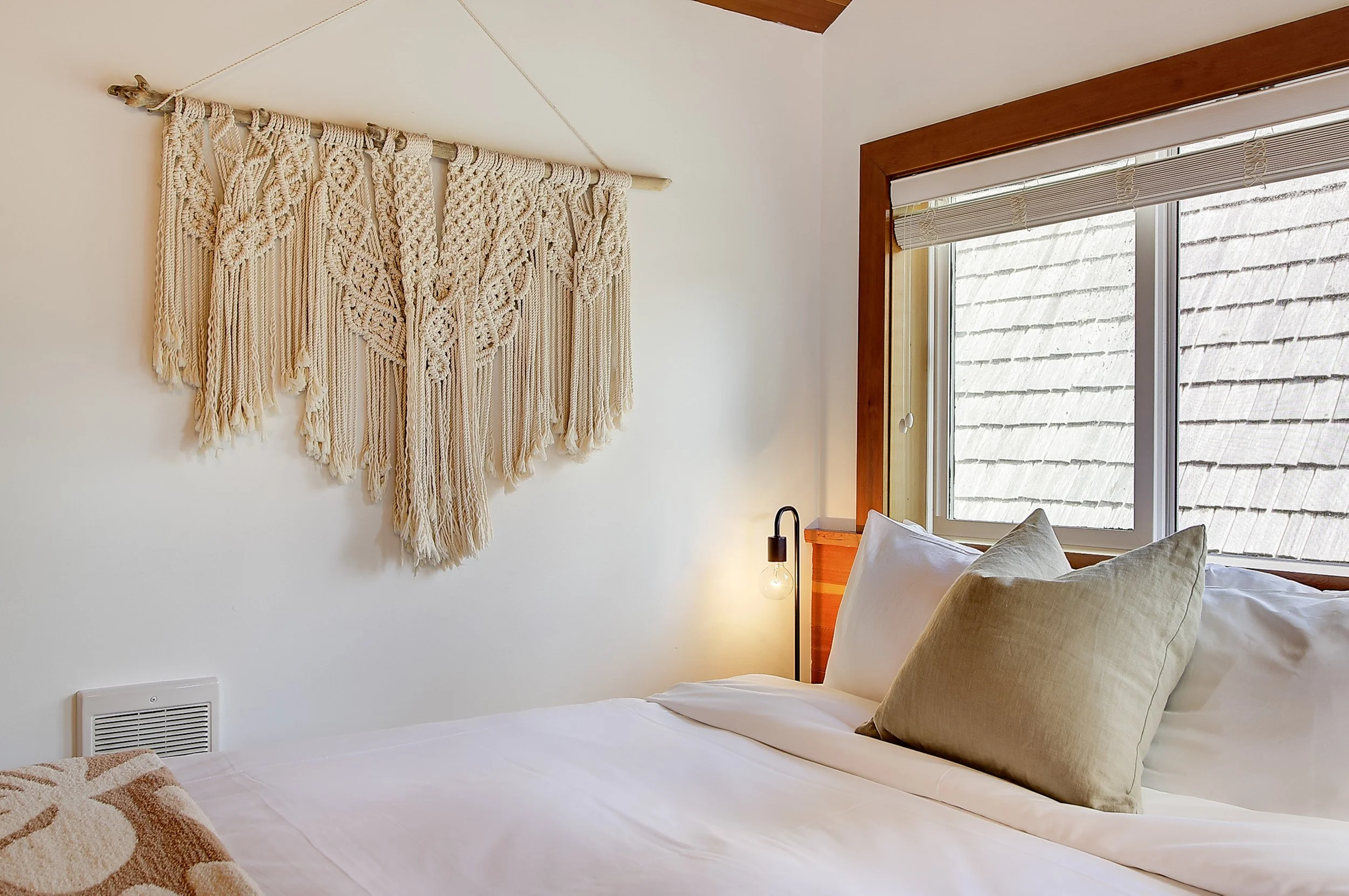 A cozy bedroom corner with a bed featuring beige and white pillows and white linens. Above the bed, a large, textured macrame wall hanging decorates the white wall. There is a wooden-framed window with partially pulled-up blinds allowing natural ligh