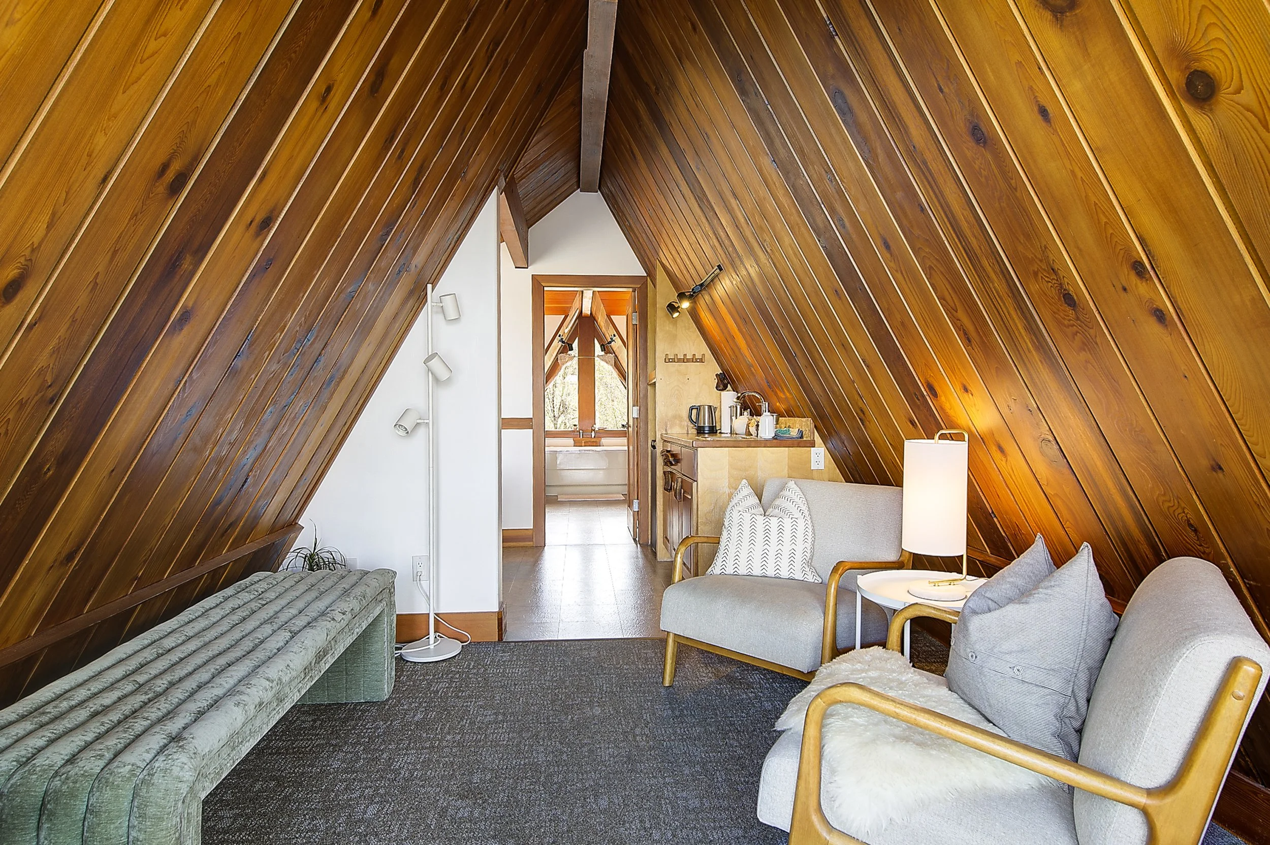An attic living space with sloped wooden paneled ceiling, furnished with chairs, a sofa, and a small table, with a view into a bathroom at the back.