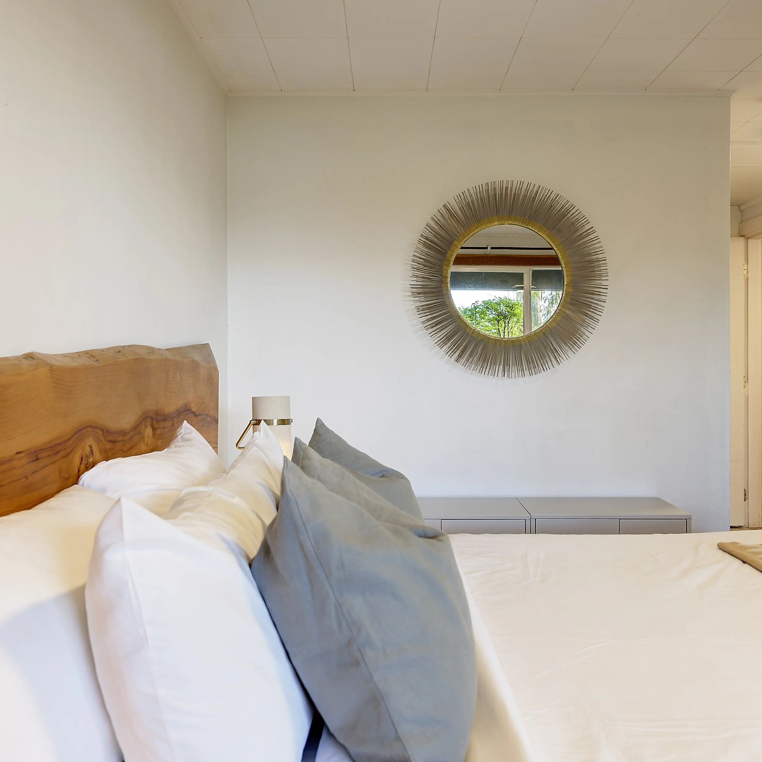 A bedroom with a wooden bed headboard, bedside lamp, and a large circular decorative mirror with a sunburst frame hanging on a white wall, with a window showing greenery outside.