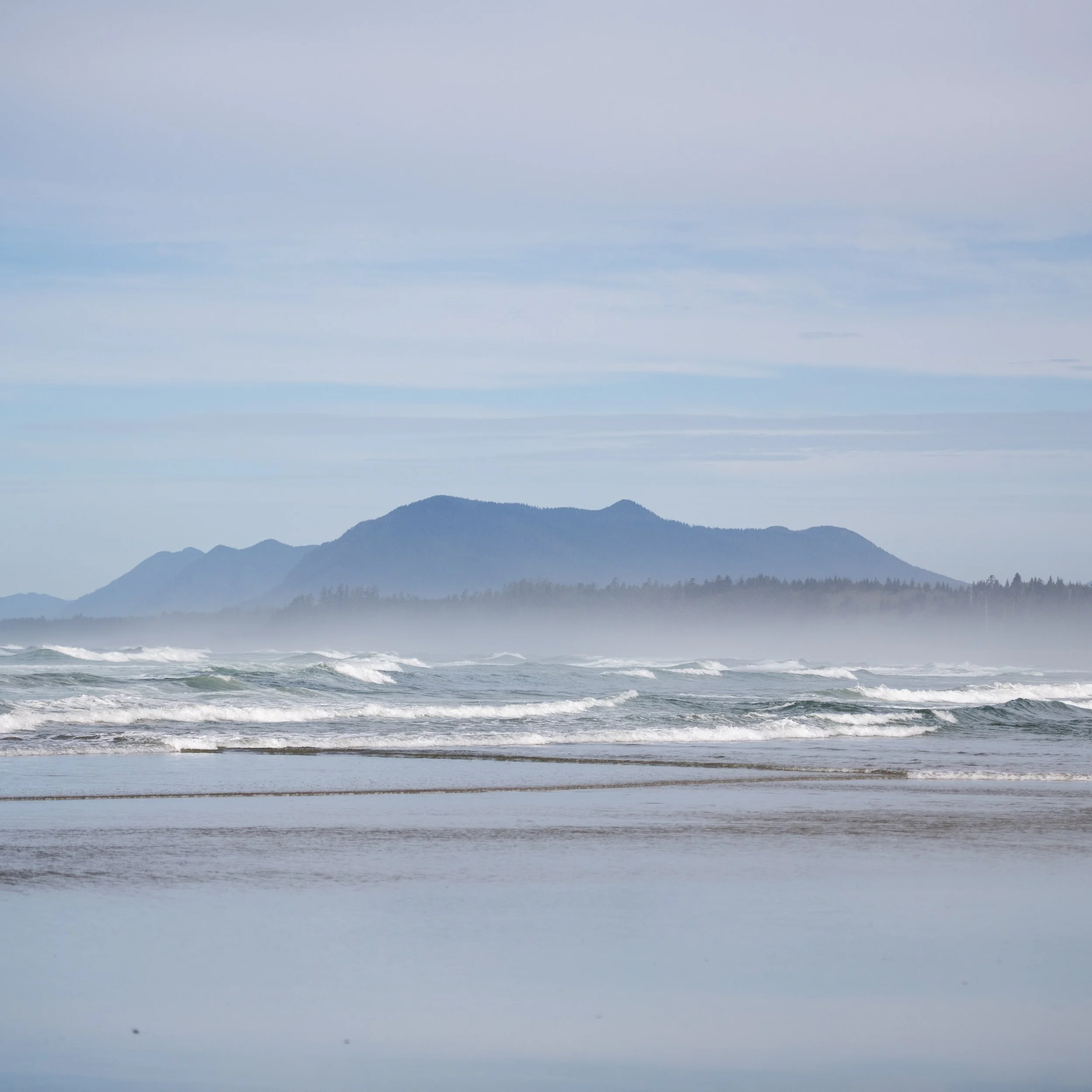 Scenic view of blue ocean waves crashing and a mountainous landscape in the distance under cloudy sky.