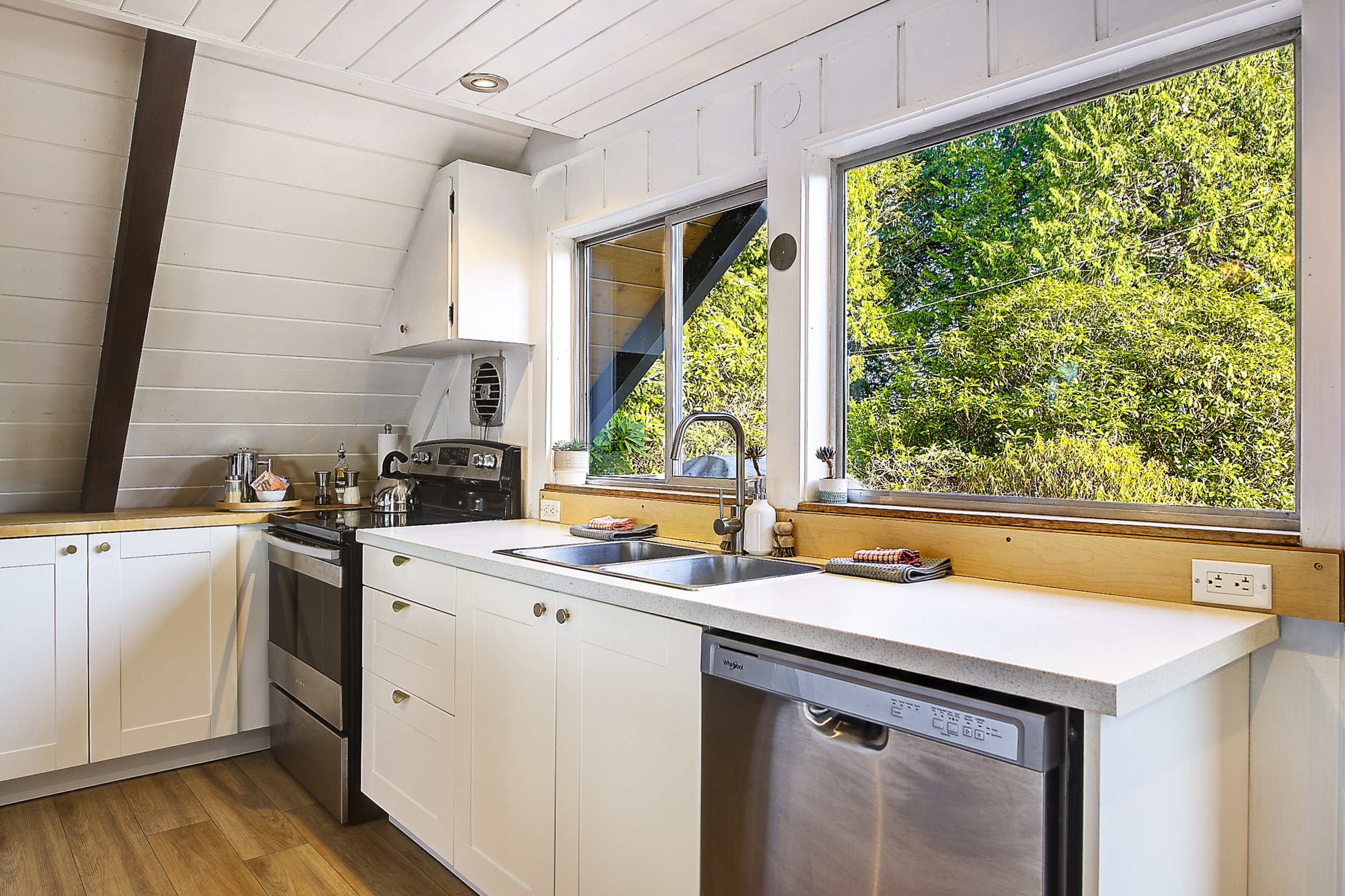 Kitchen with white cabinets, stainless steel oven, dishwasher, ample counter space, and a large window showing green leafy trees outside.