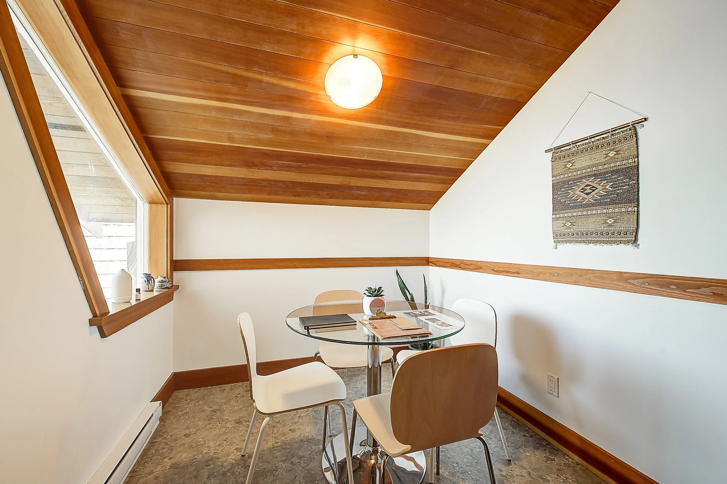 A small round glass dining table with four chairs, two white and two wood, in a room with white walls and wood-paneled sloped ceiling. The table has some books, magazines, and a potted plant. A rectangular woven wall hanging is on the right wall, and