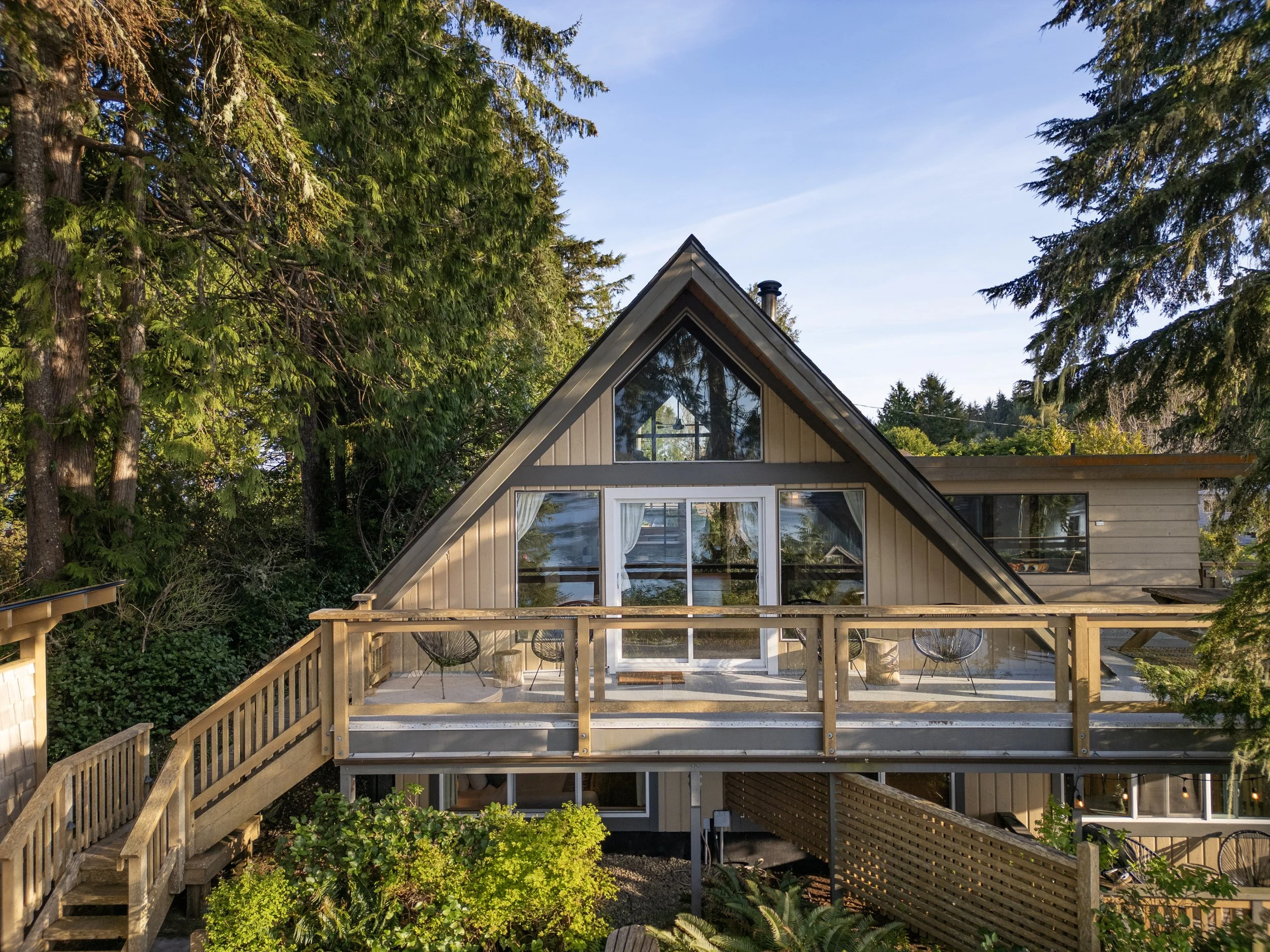 A house with a steep triangular roof and large windows surrounded by tall trees, with a wooden deck and railing in the foreground, set on a clear day with blue sky.